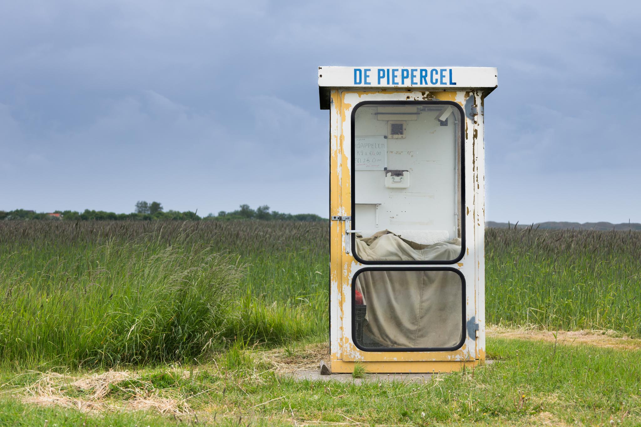 Weathered yellow phone booth labeled De Piepercel in grassy field under cloudy sky.
