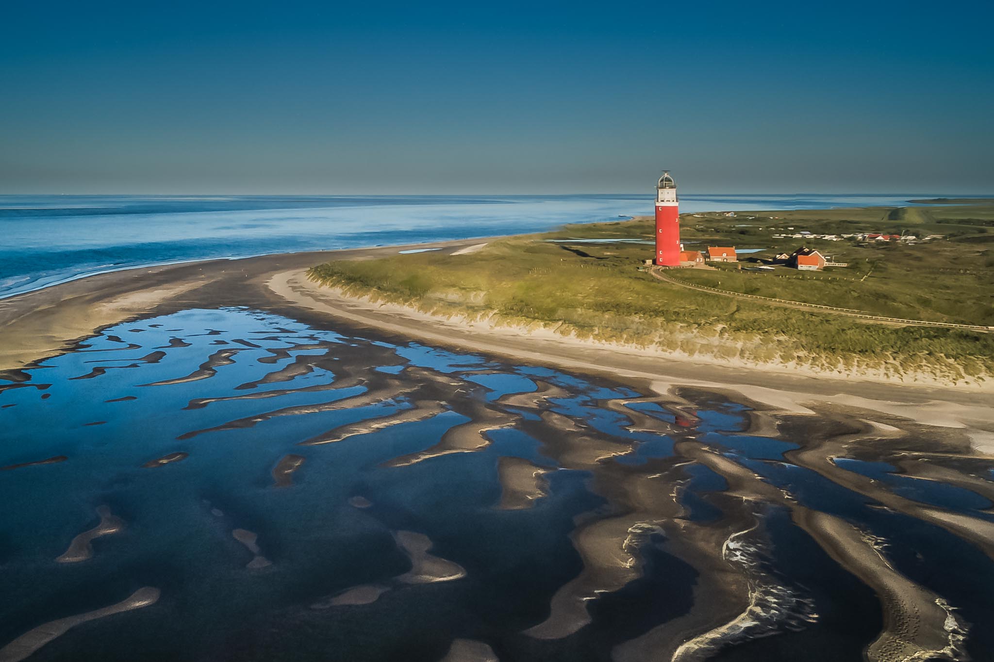 Aerial view of a red lighthouse on a coastal landscape with sandy beach and tidal pools under a clear blue sky.