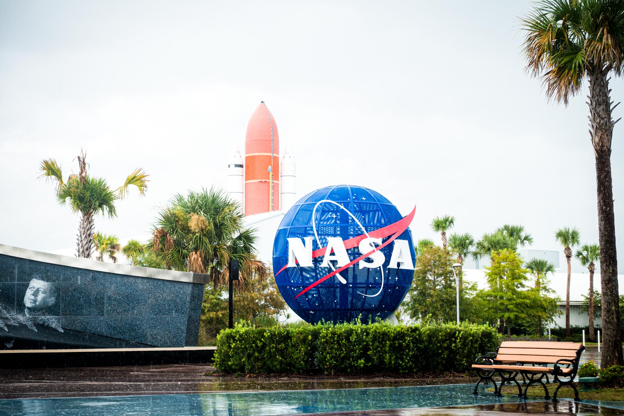 NASA globe and rocket display with palm trees and a bench in a green park setting.