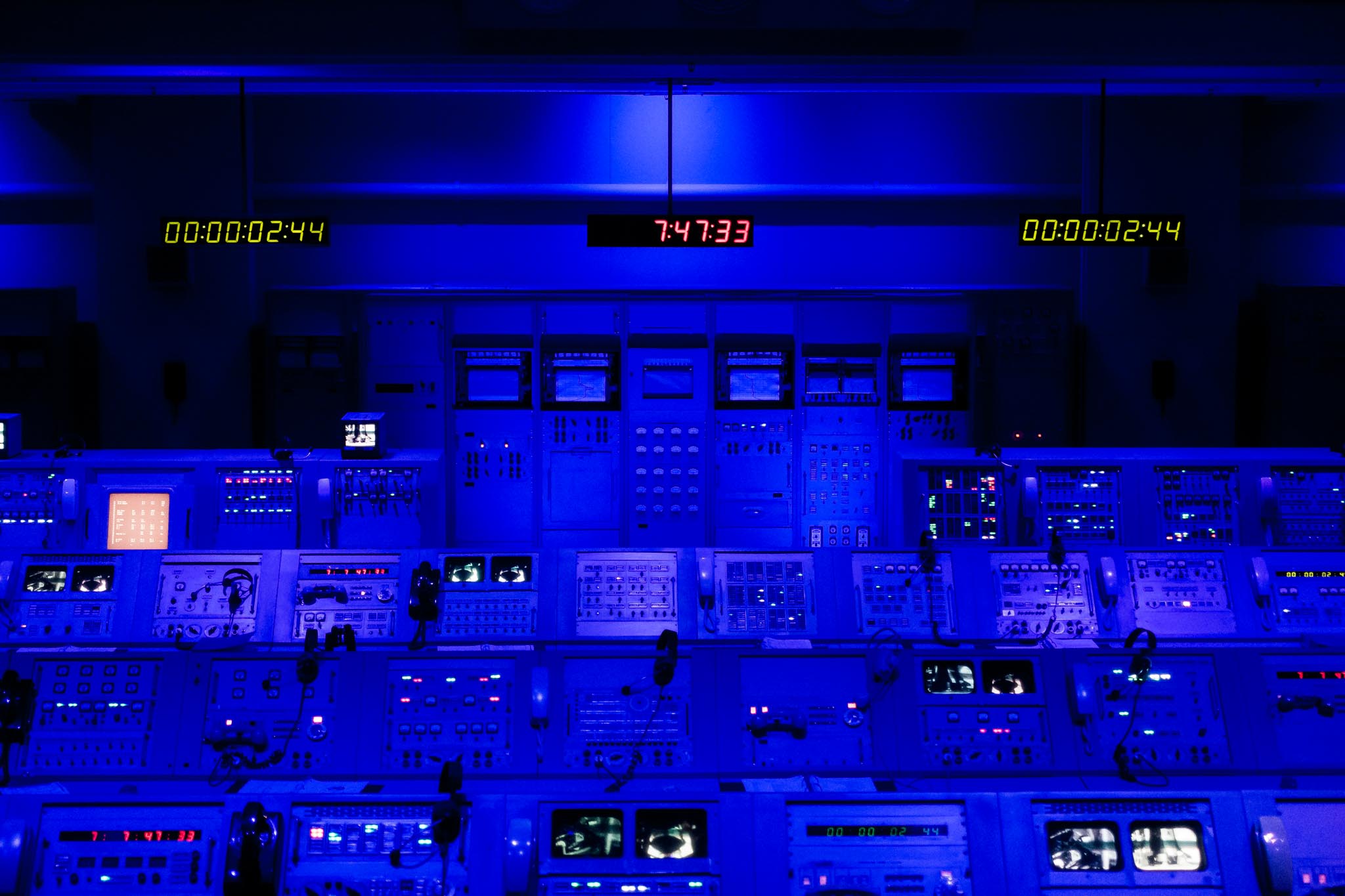 Blue-lit control room with multiple illuminated panels and digital clocks displaying time.