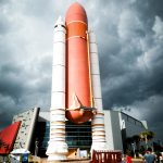 Space shuttle replica at Kennedy Space Center with dramatic cloudy sky, visitors at entrance.
