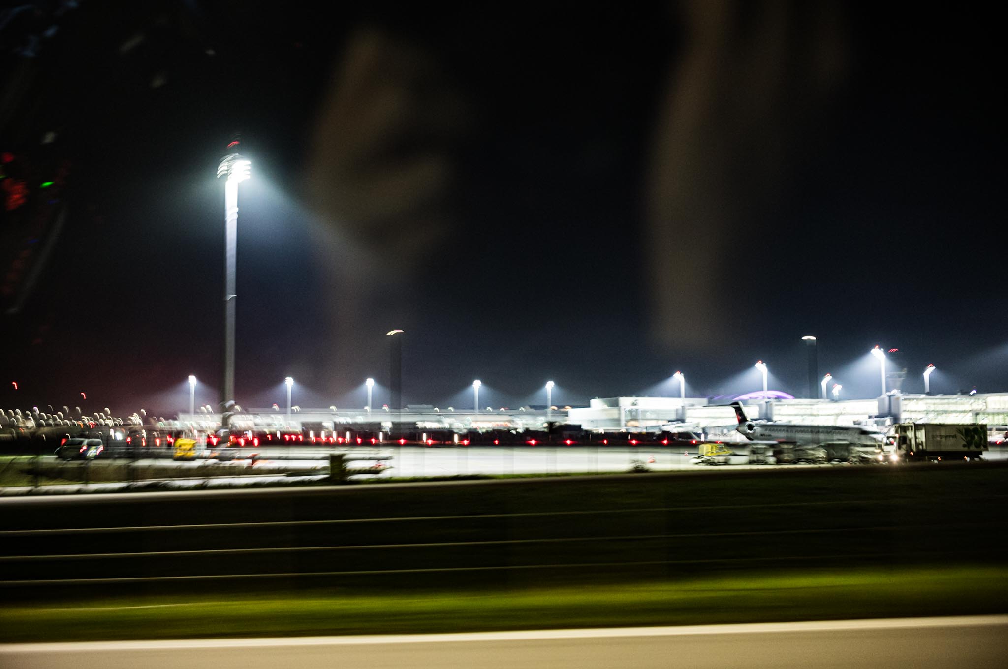 Illuminated airport runway at night with airplanes and vehicles visible, under tall glowing light posts.