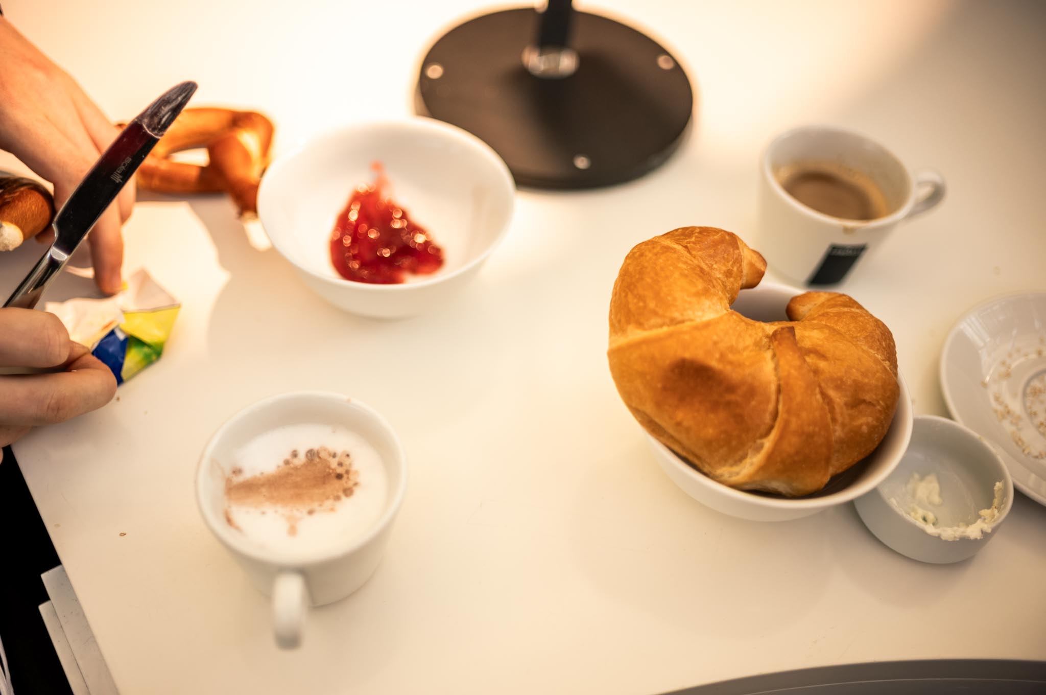 Breakfast setup with croissant, bowl of jam, pretzel, and cups of coffee and cappuccino on a white table.