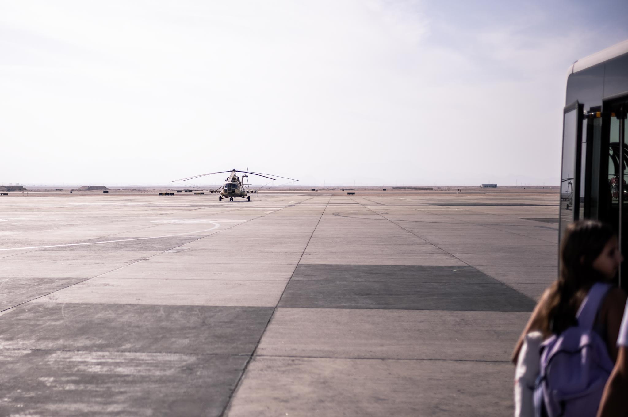 Helicopter on airport tarmac with bus and person in foreground under a cloudy sky.