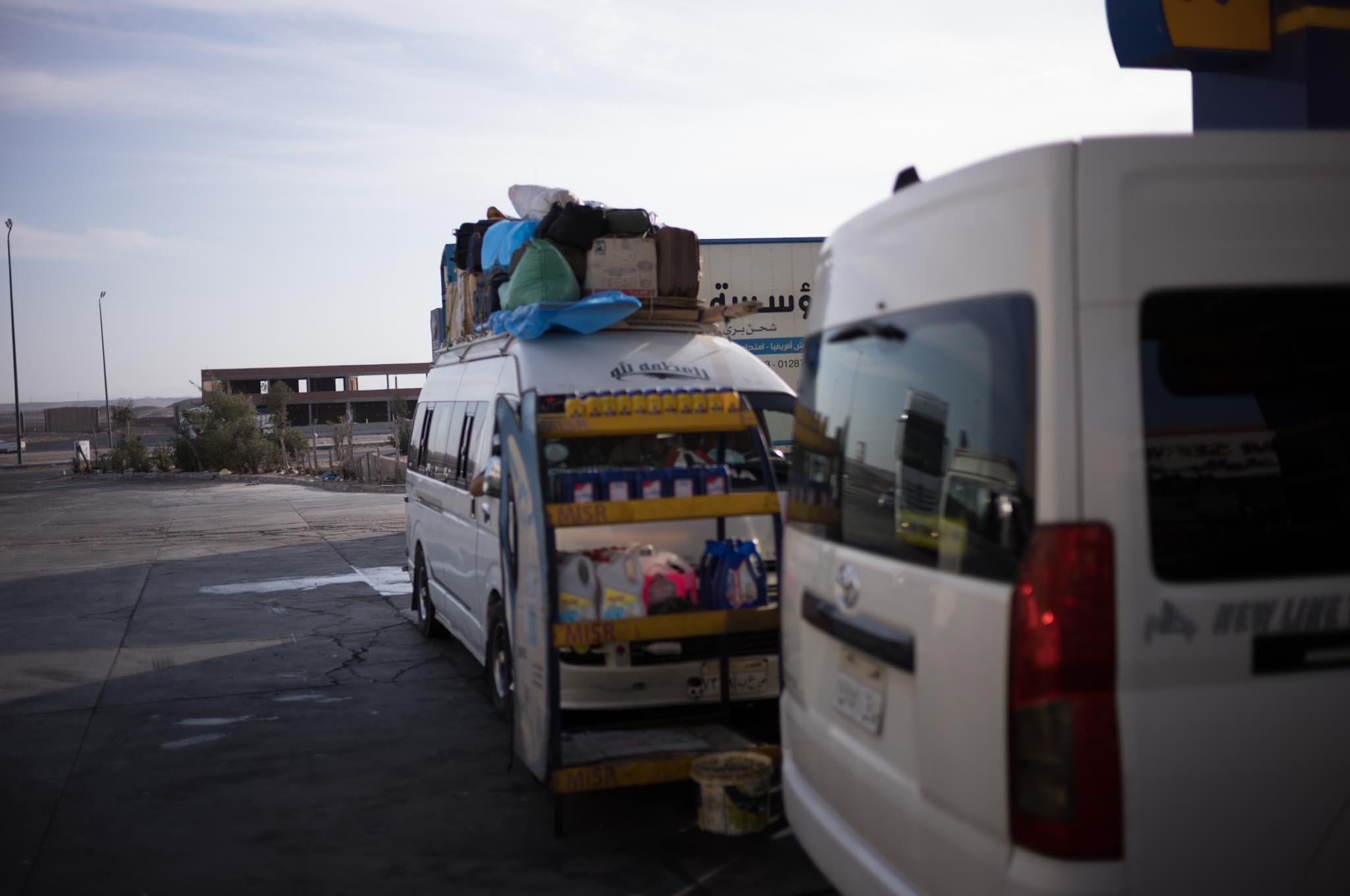 White vans parked at a gas station, one with a rooftop loaded with bags and an open side showcasing shelves with goods.