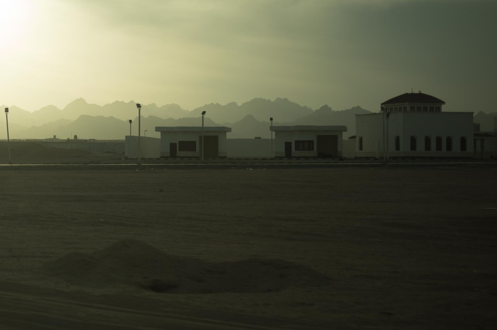 Desert landscape at sunset with white buildings and distant mountains silhouetted against a hazy sky.