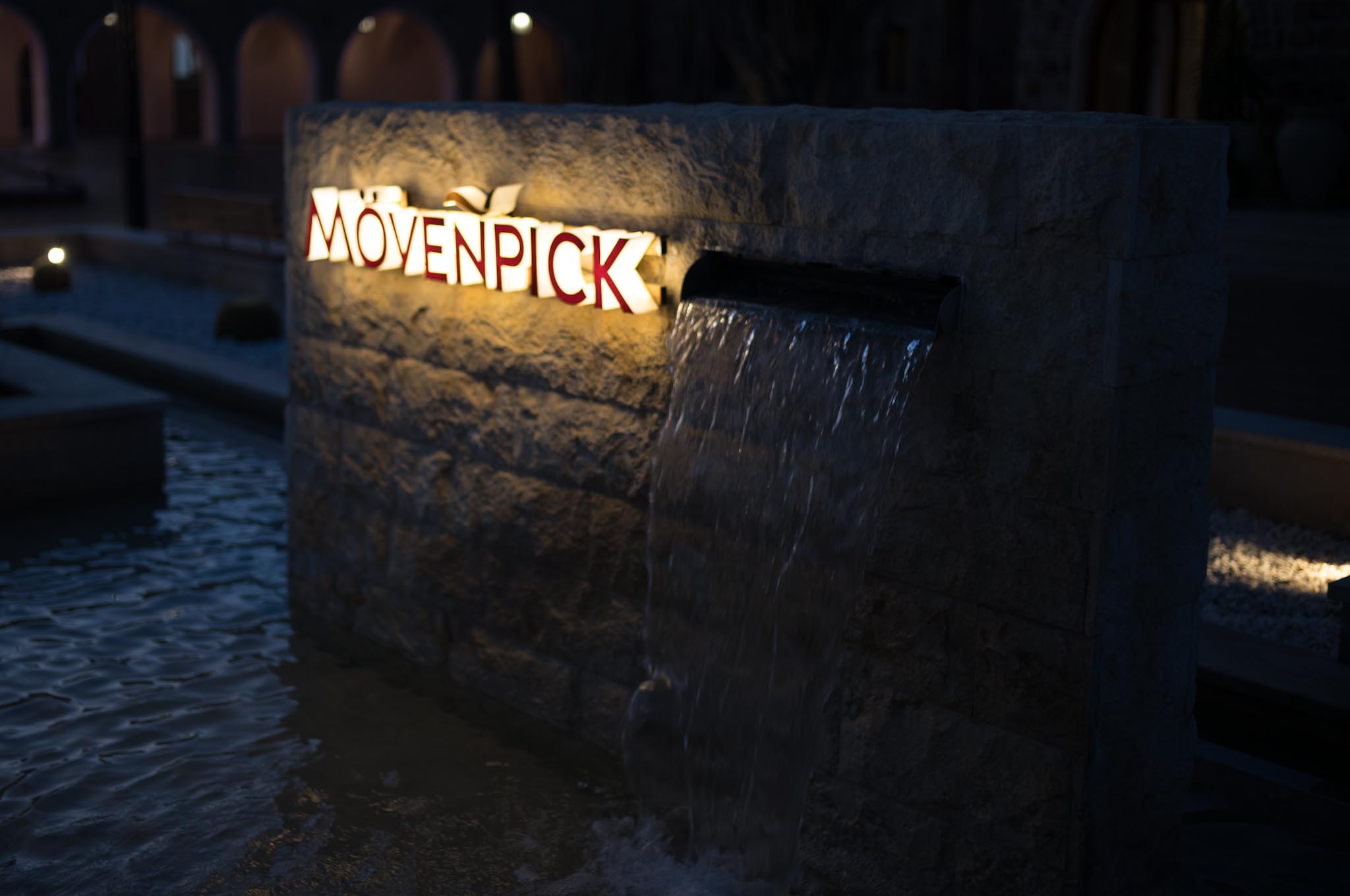 Illuminated Mövenpick sign on a stone fountain with cascading water at night.