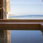 Ocean view from a relaxing poolside terrace with wooden shutters and straw umbrella, overlooking a calm, sunny beach.