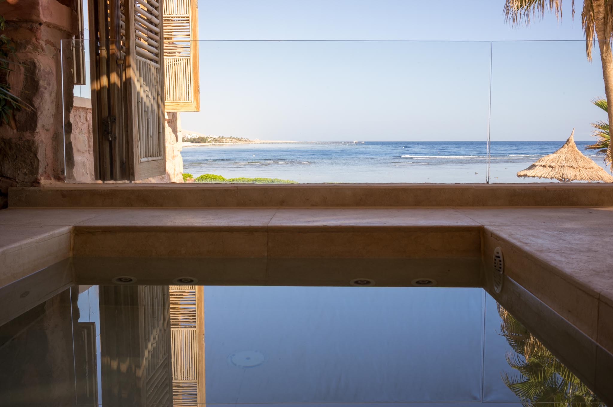 Ocean view from a relaxing poolside terrace with wooden shutters and straw umbrella, overlooking a calm, sunny beach.
