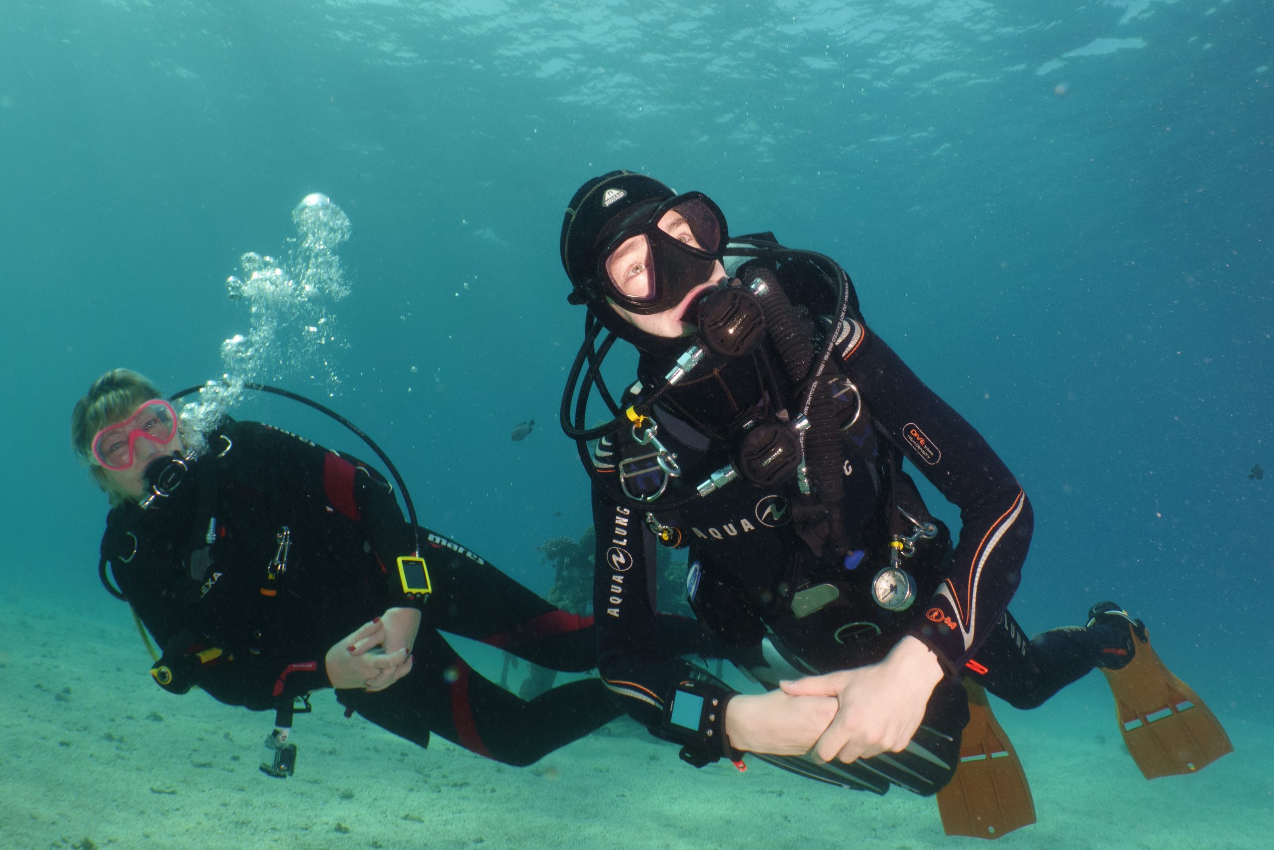 Two scuba divers in wetsuits and gear exploring an underwater scene with clear blue water.