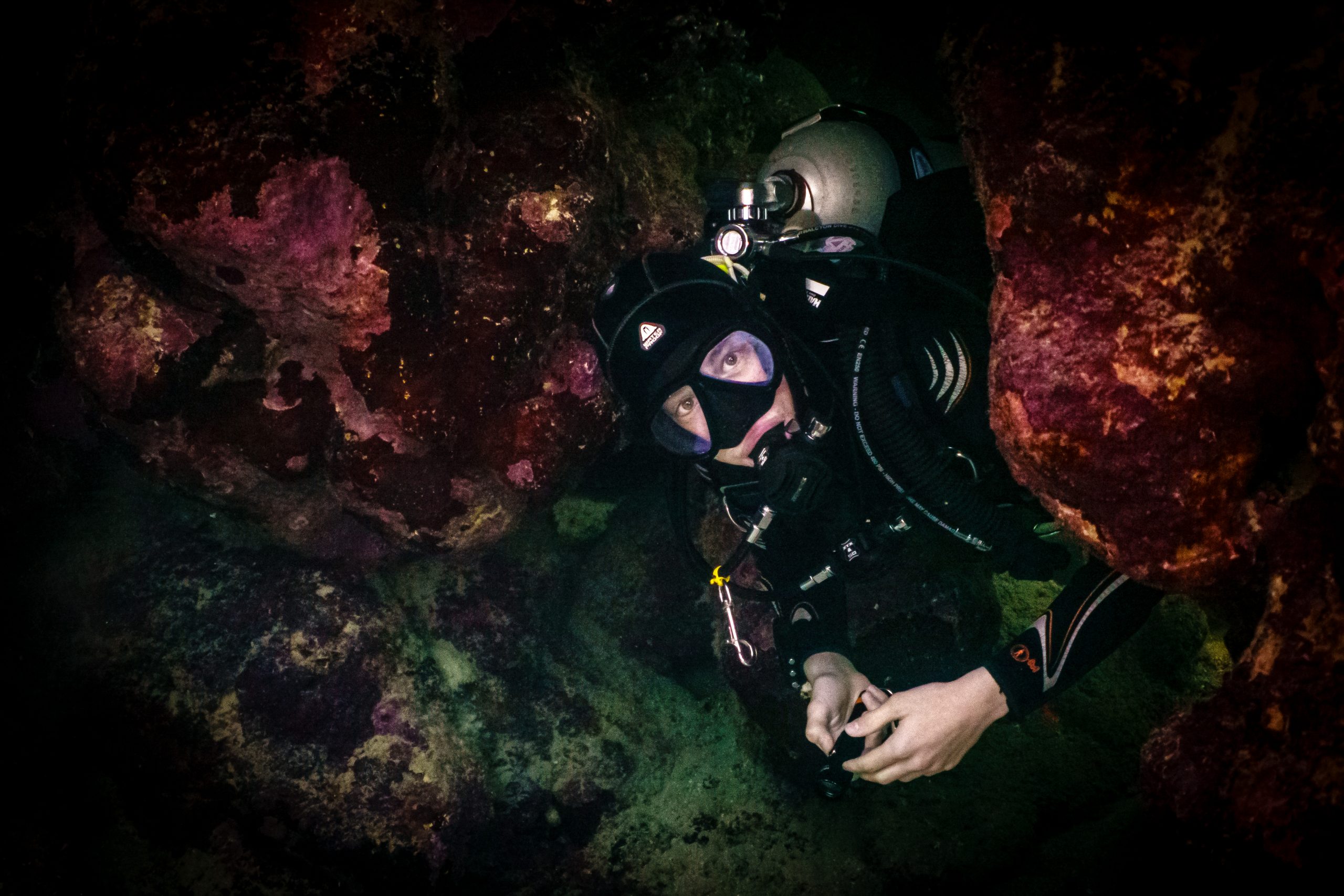 Scuba diver exploring underwater cave with rocky walls and vibrant coral formations.
