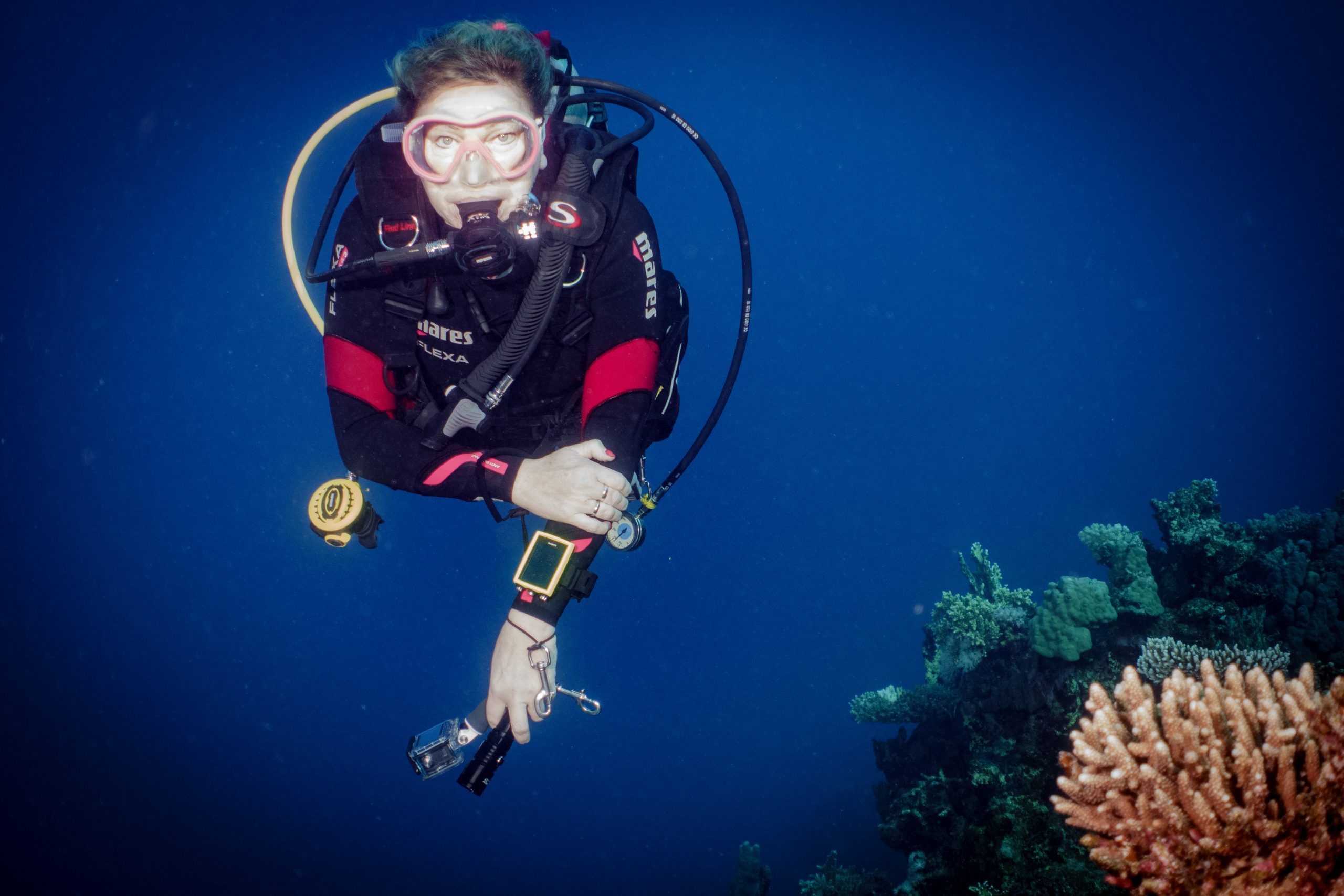 Scuba diver exploring coral reef underwater, wearing red and black gear with a camera in hand.