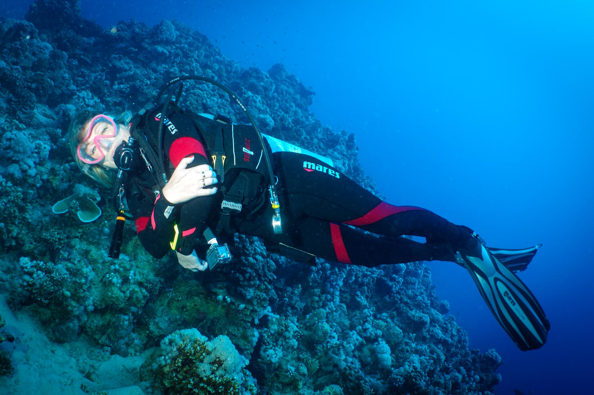 Scuba diver in wetsuit exploring vibrant coral reef underwater, holding camera in clear blue ocean.