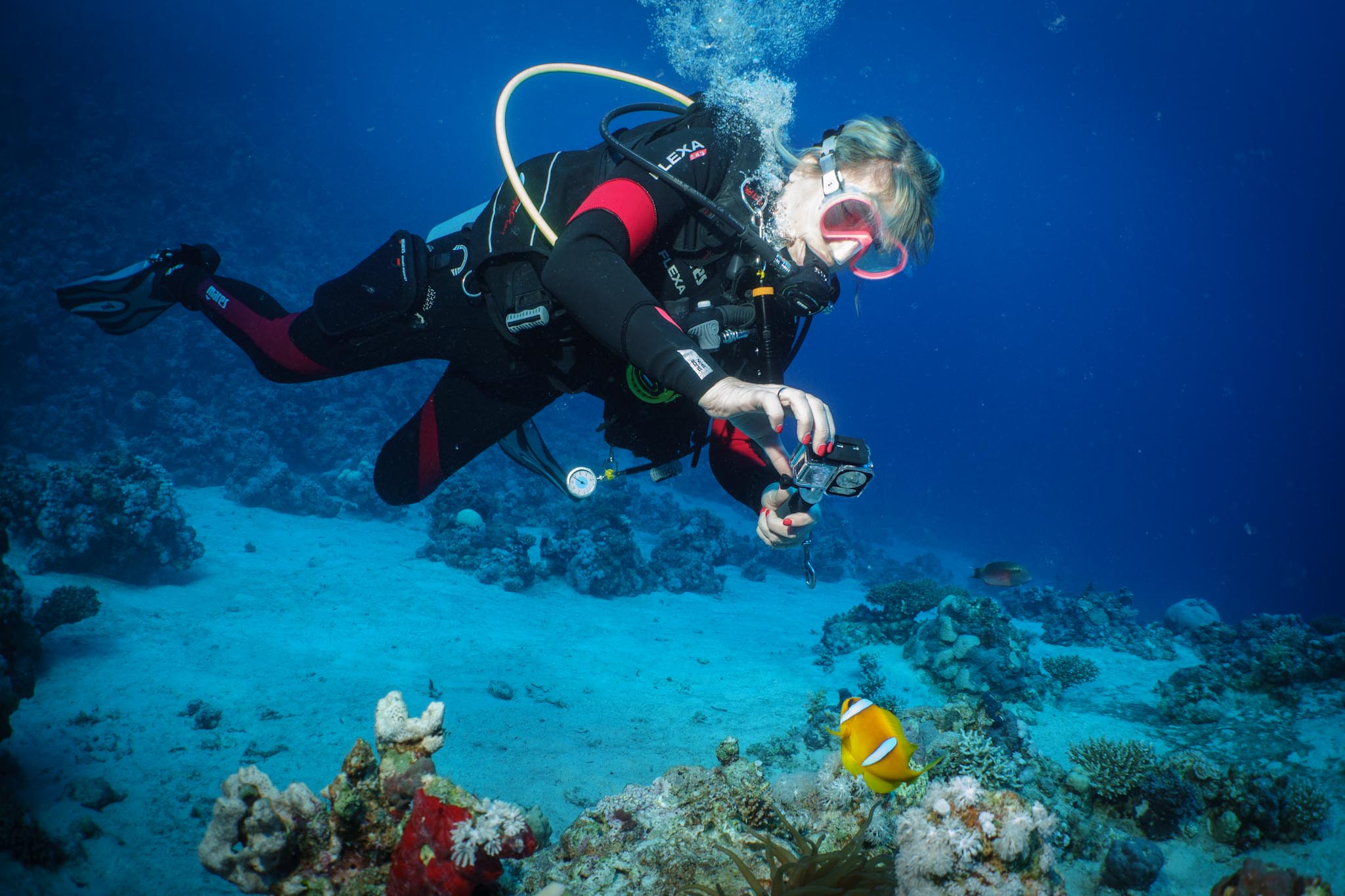 Scuba diver photographing a clownfish on a vibrant coral reef under clear blue ocean water.