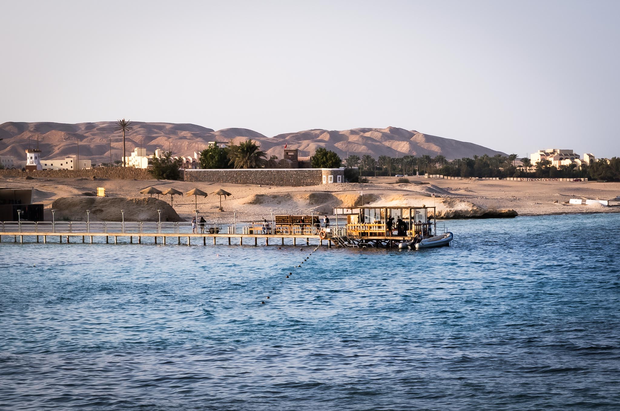 Scenic coastal view with pier extending into turquoise waters, against backdrop of desert landscape and buildings.