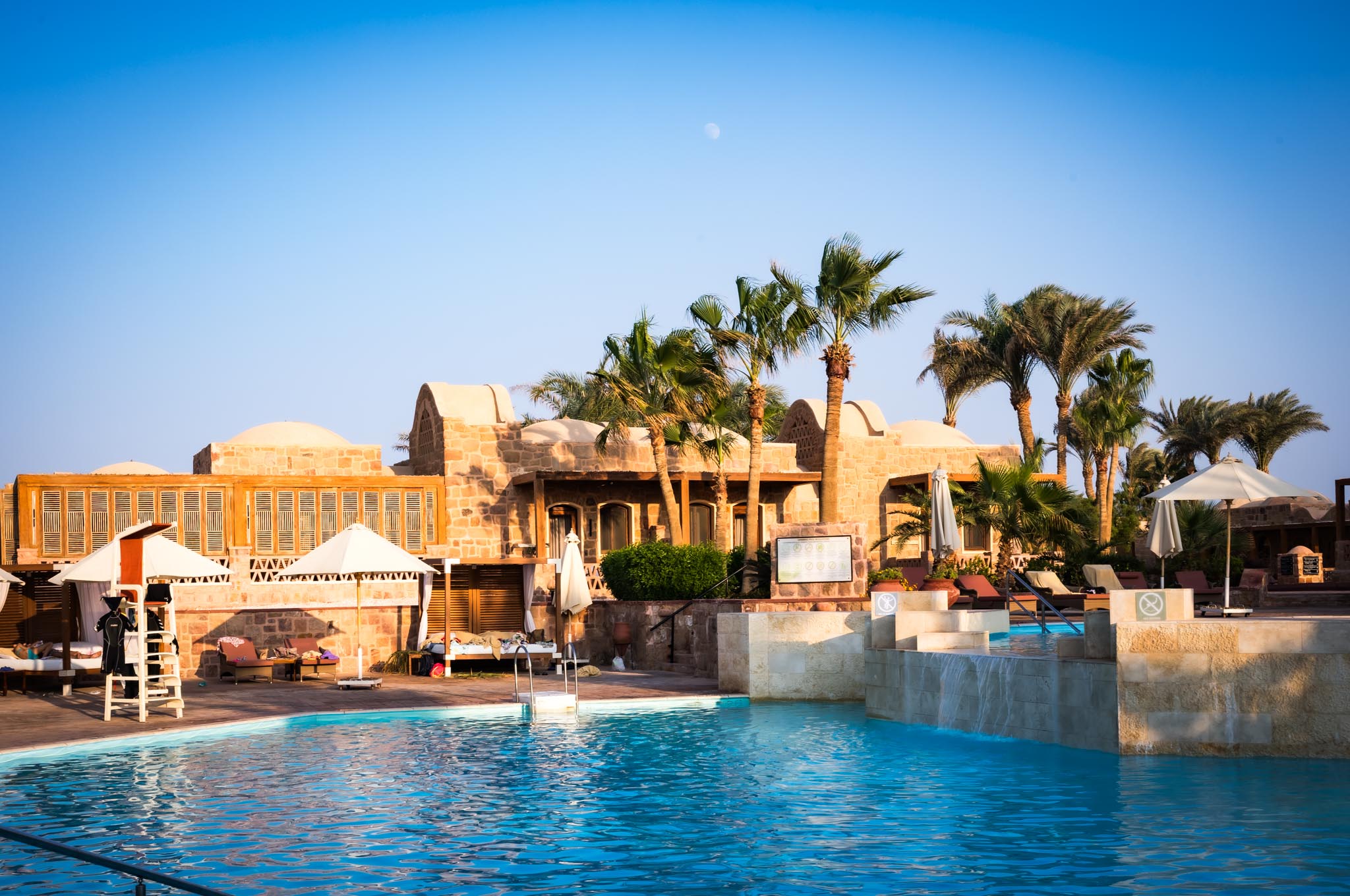 Luxury resort pool with loungers, umbrellas, palm trees, and stone buildings under a clear blue sky.