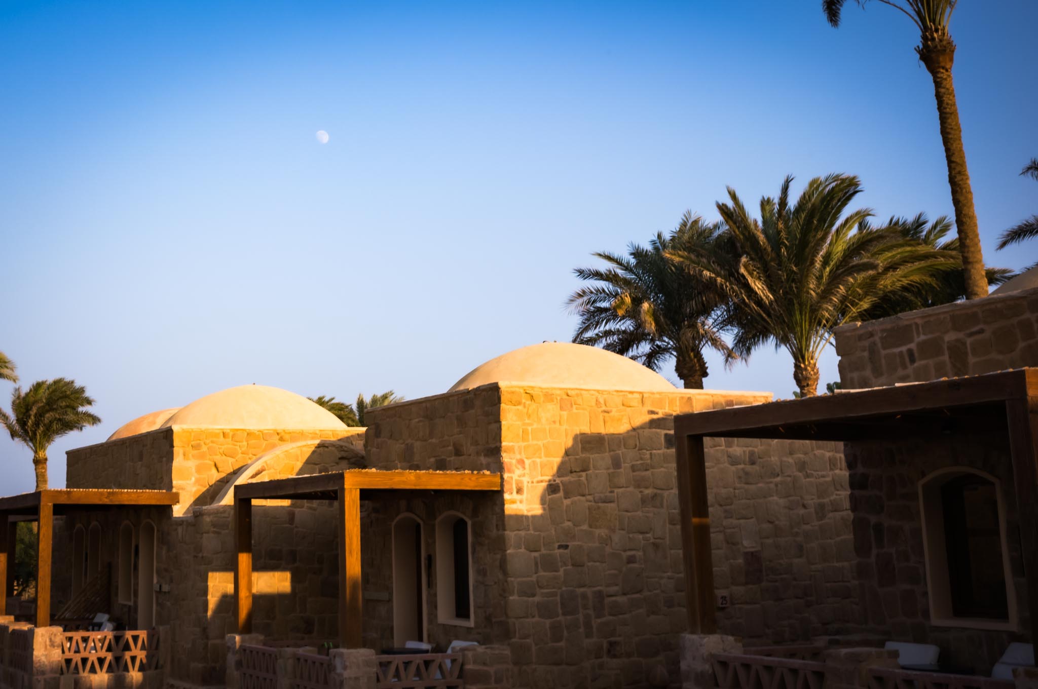Stone buildings with domed roofs and palm trees under a blue sky with visible moon.