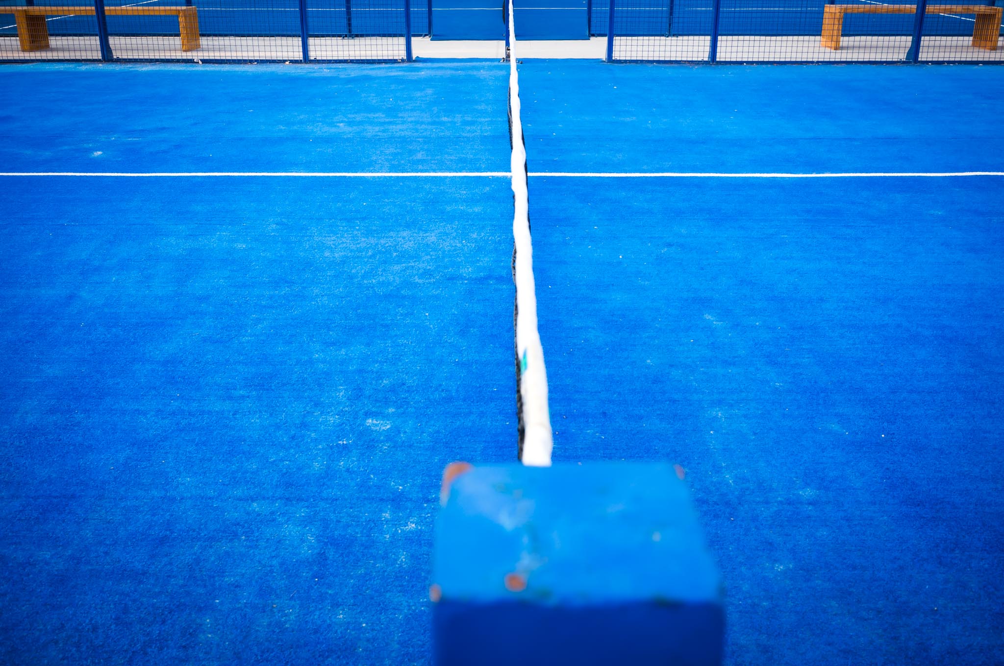 Blue paddle tennis court with a white line and net dividing the vibrant blue surface, surrounded by a fence and benches.
