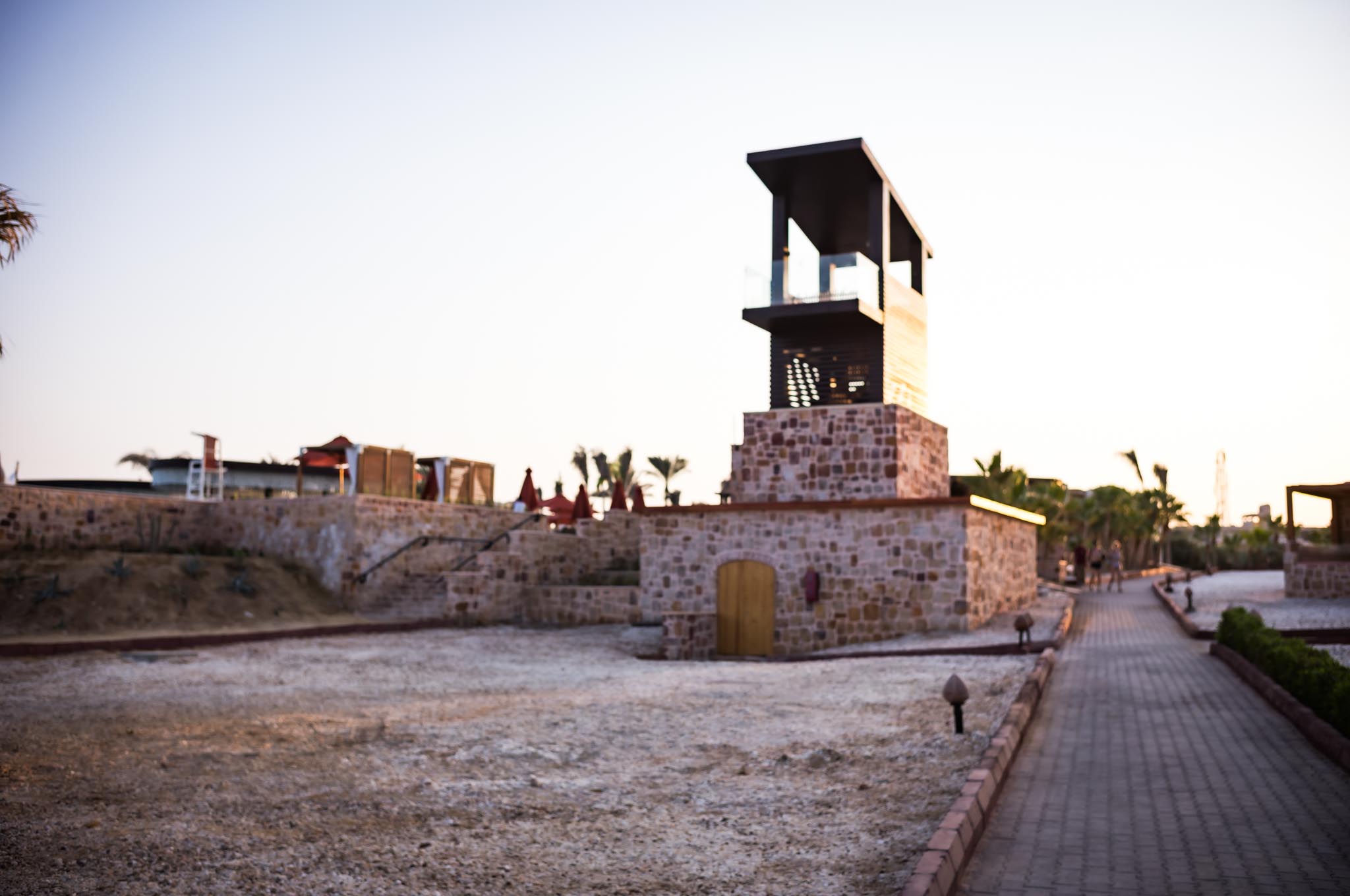 Stone building with modern tower, surrounded by palm trees at sunset, on a paved walkway in an open area.