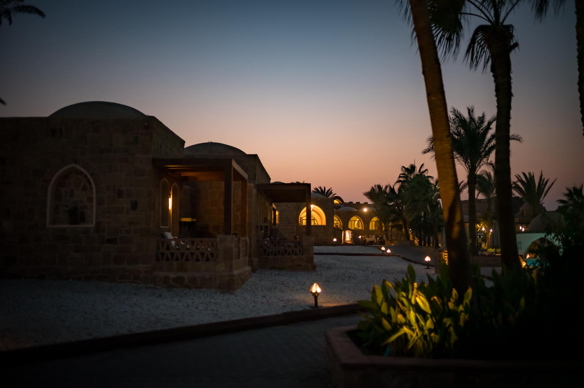 Sunset view of a rustic stone villa with illuminated windows, surrounded by palm trees and warm twilight sky.