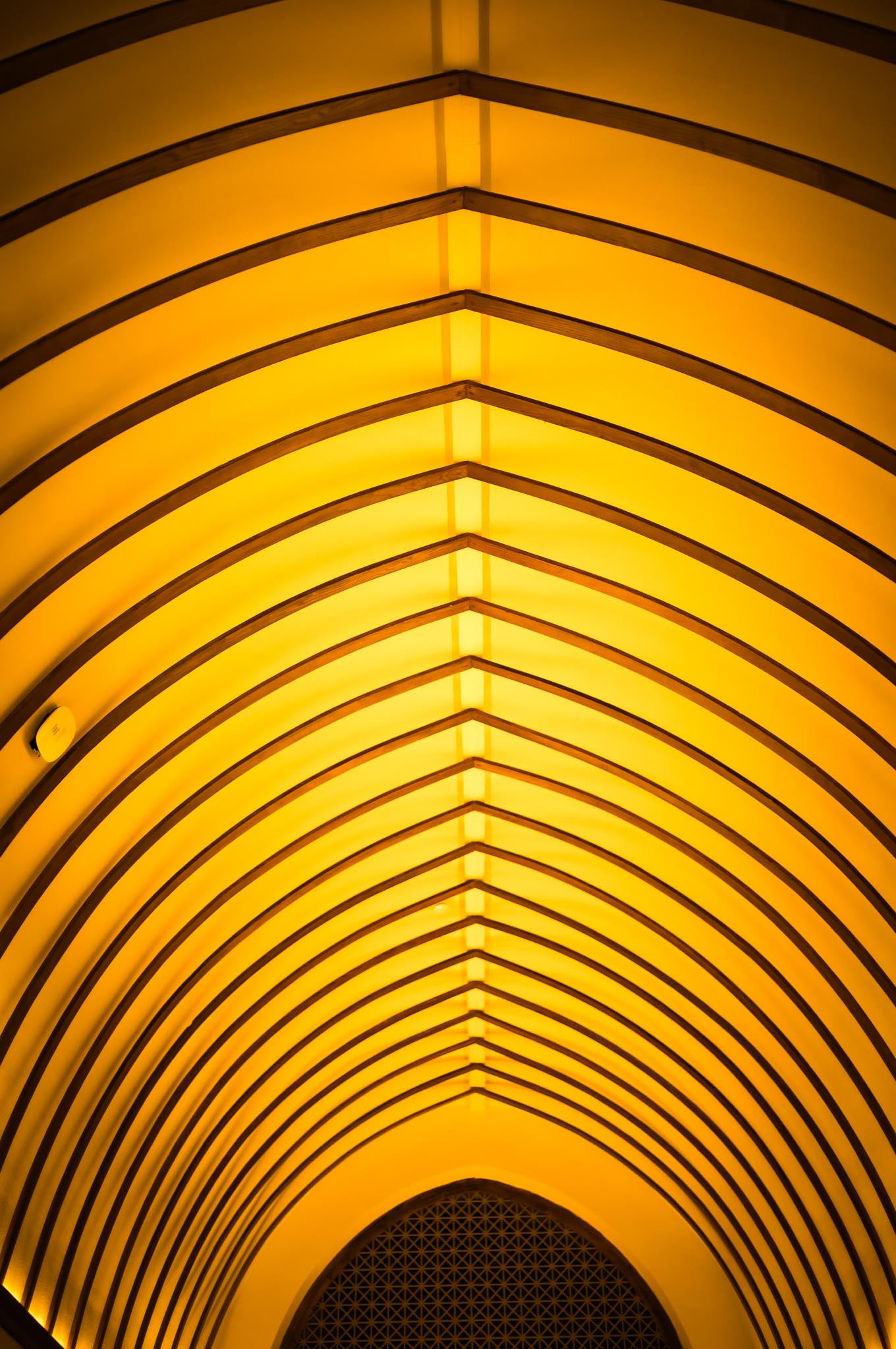 Illuminated yellow ceiling with arched wooden beams in geometric pattern, creating a modern architectural design.