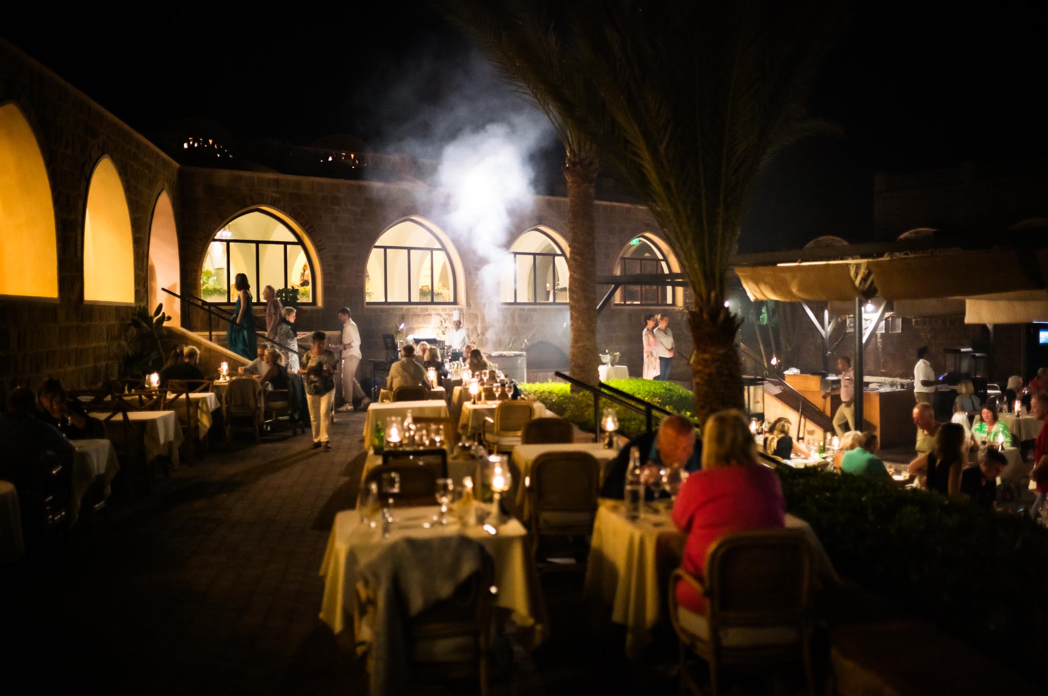Outdoor evening restaurant with people dining, candle-lit tables, stone arches, and ambient lighting creating a cozy atmosphere.