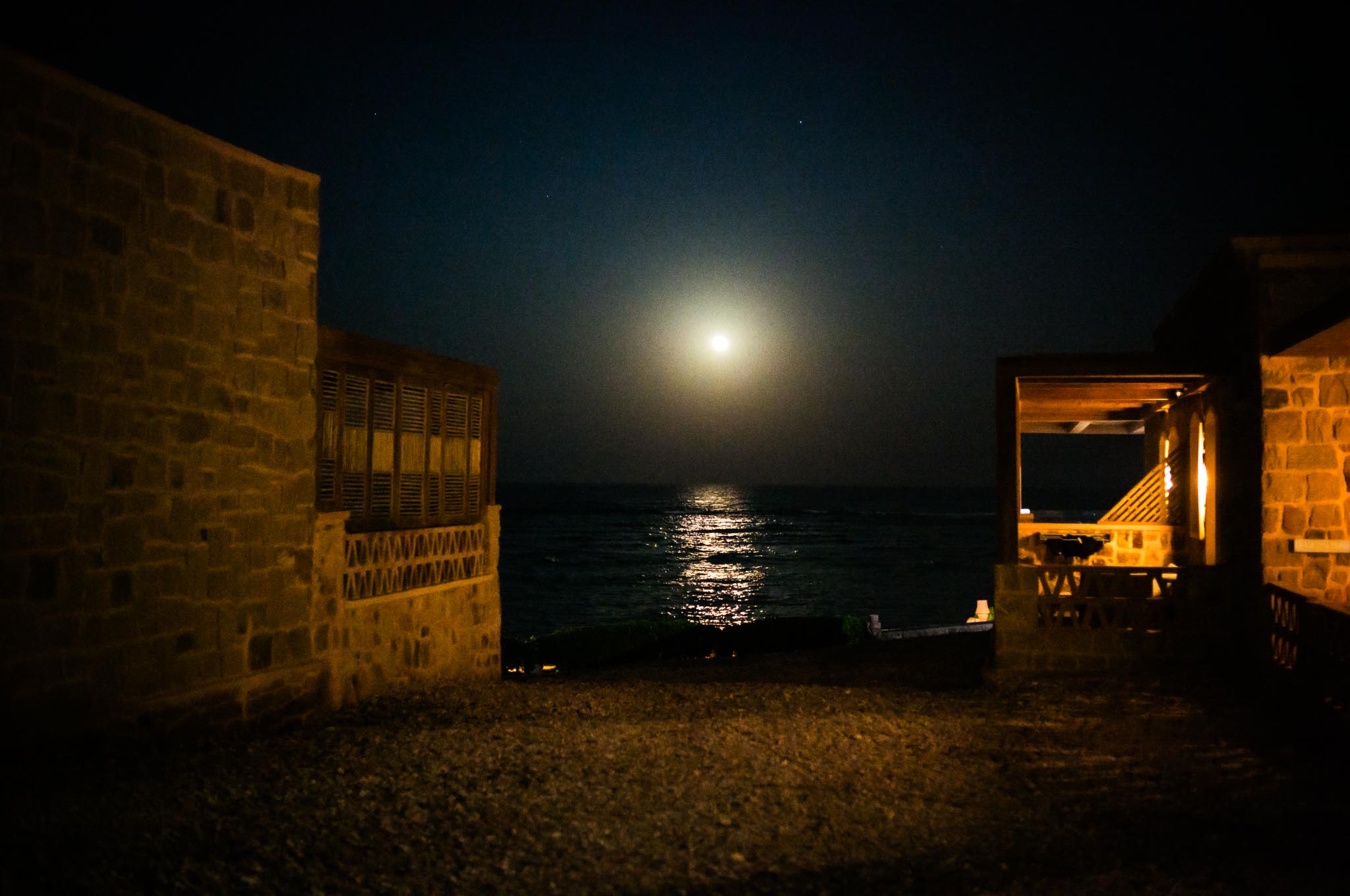Moonlit ocean view framed by rustic stone buildings at night.