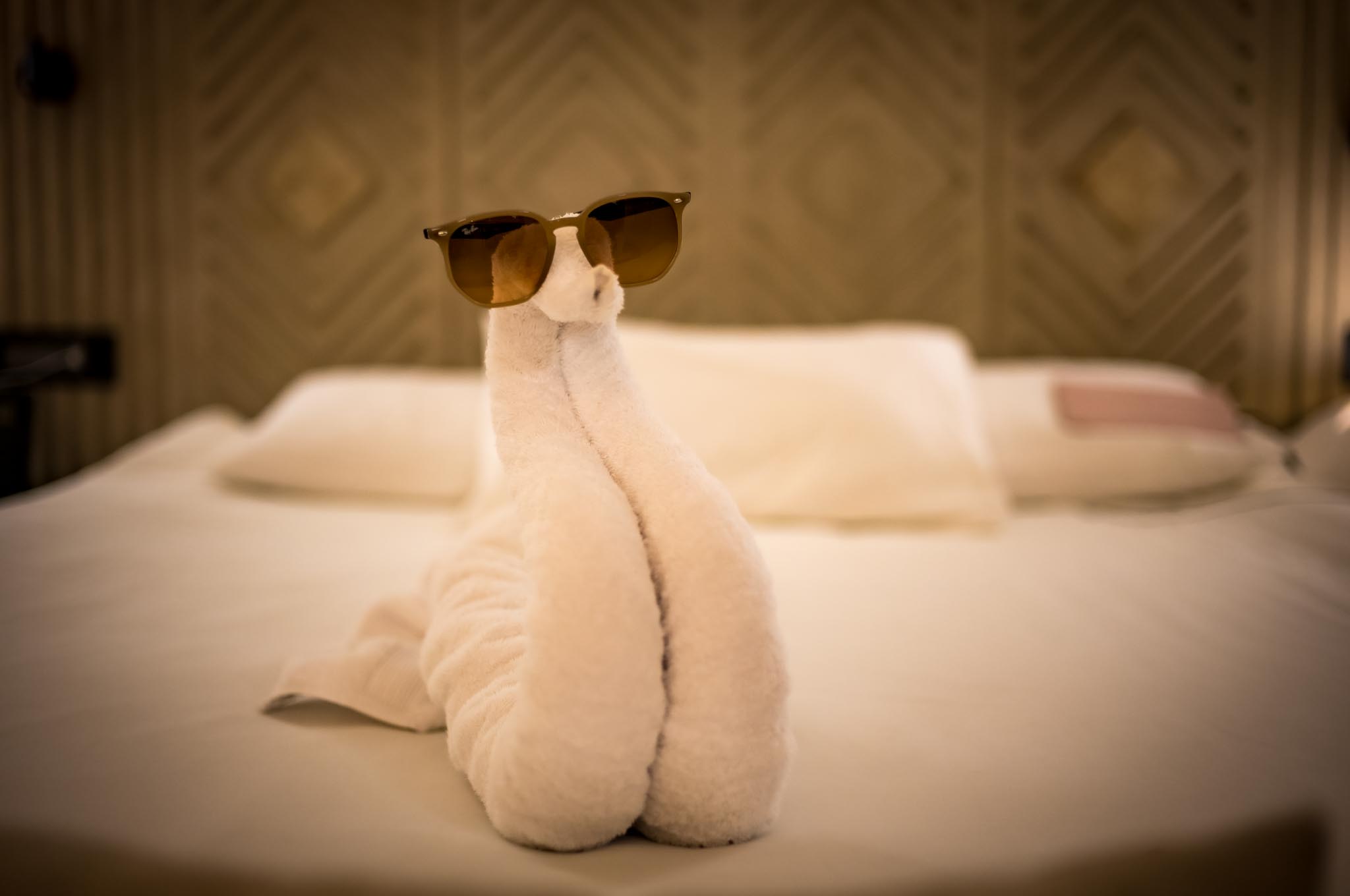 Towel swan with sunglasses sits on a neatly made bed in a hotel room.
