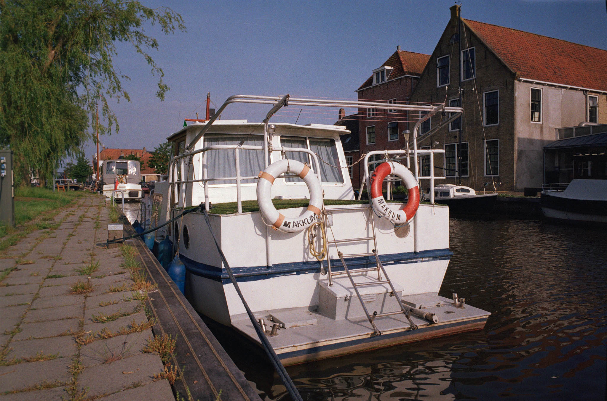 Boat docked by canal with lifebuoys labeled Makkum, surrounded by rustic buildings and trees under a clear sky.