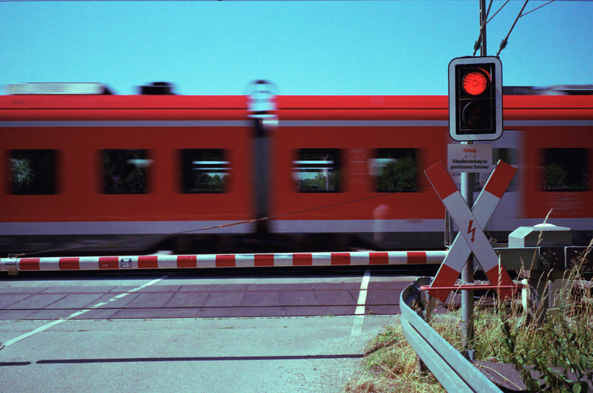 Red train passing a crossing with a closed barrier and a red traffic light under a clear blue sky.