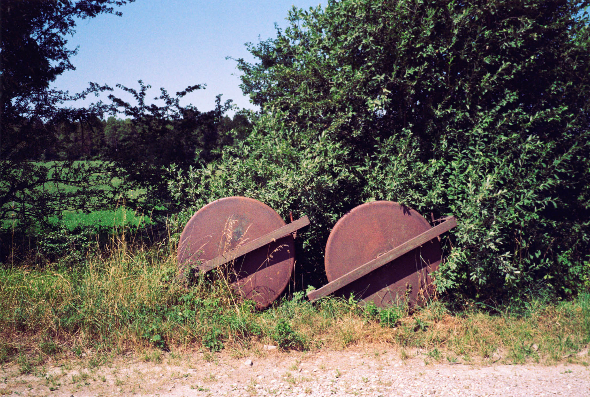 Two rusty grinding wheels leaning against overgrown bushes beside a gravel path under a clear blue sky.
