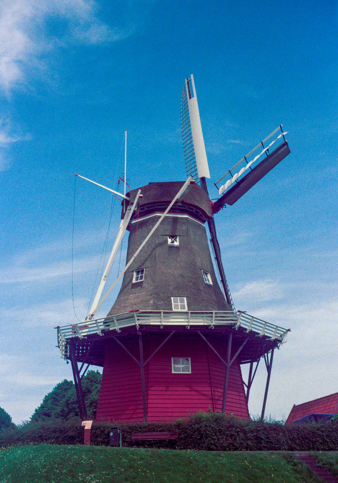 Historic red windmill with sails under a bright blue sky, surrounded by greenery.