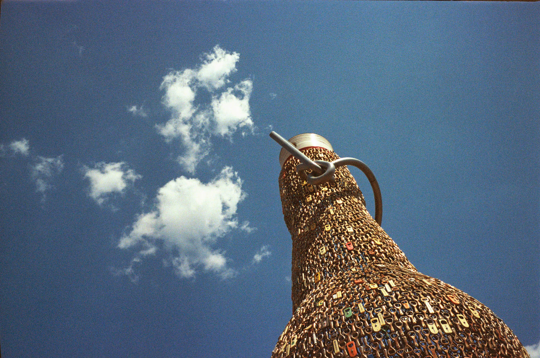 Sculpture of teapot made from zippers against a bright blue sky with scattered clouds.