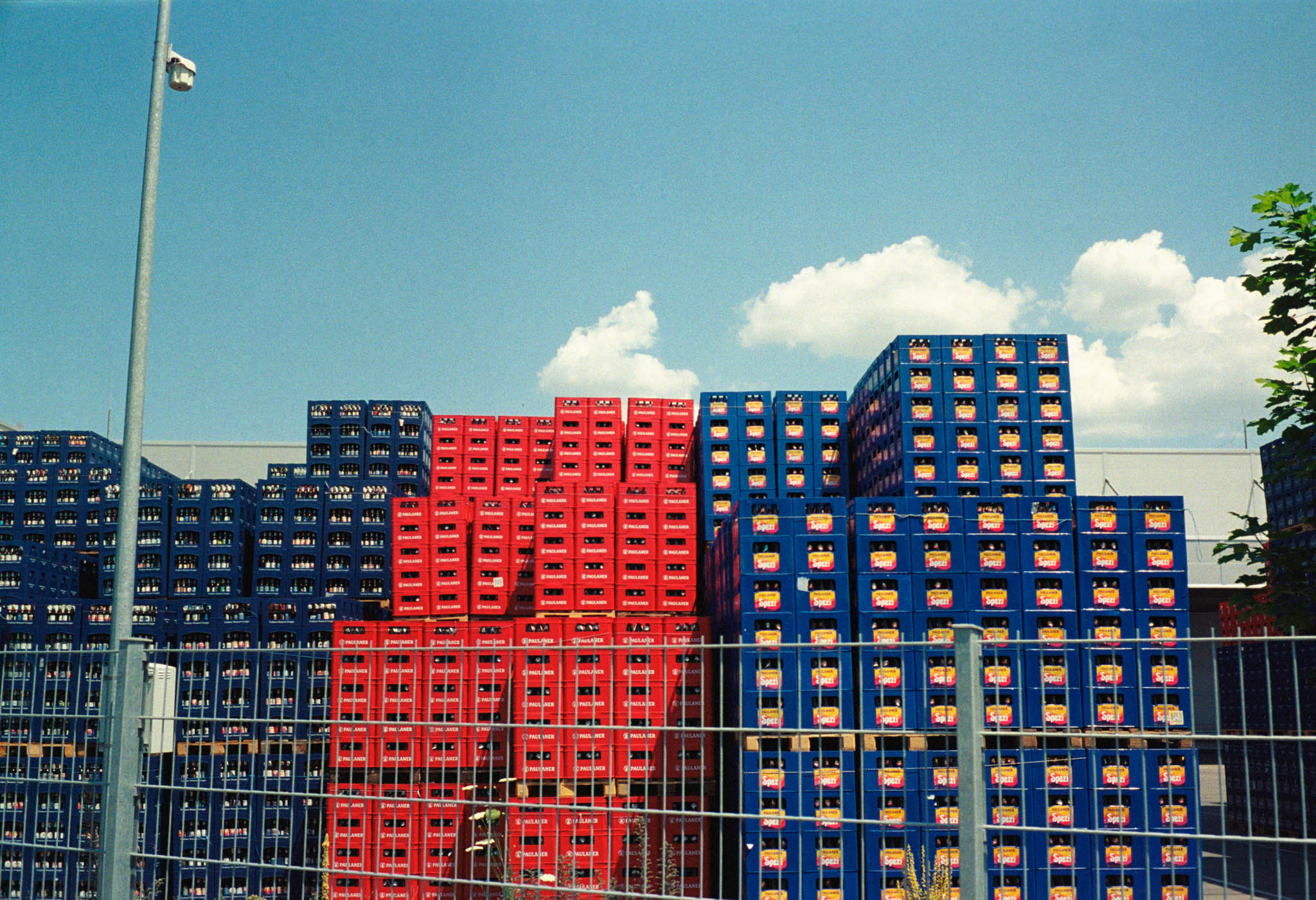 Stacks of red and blue beverage crates behind a metal fence under a clear blue sky with scattered clouds.