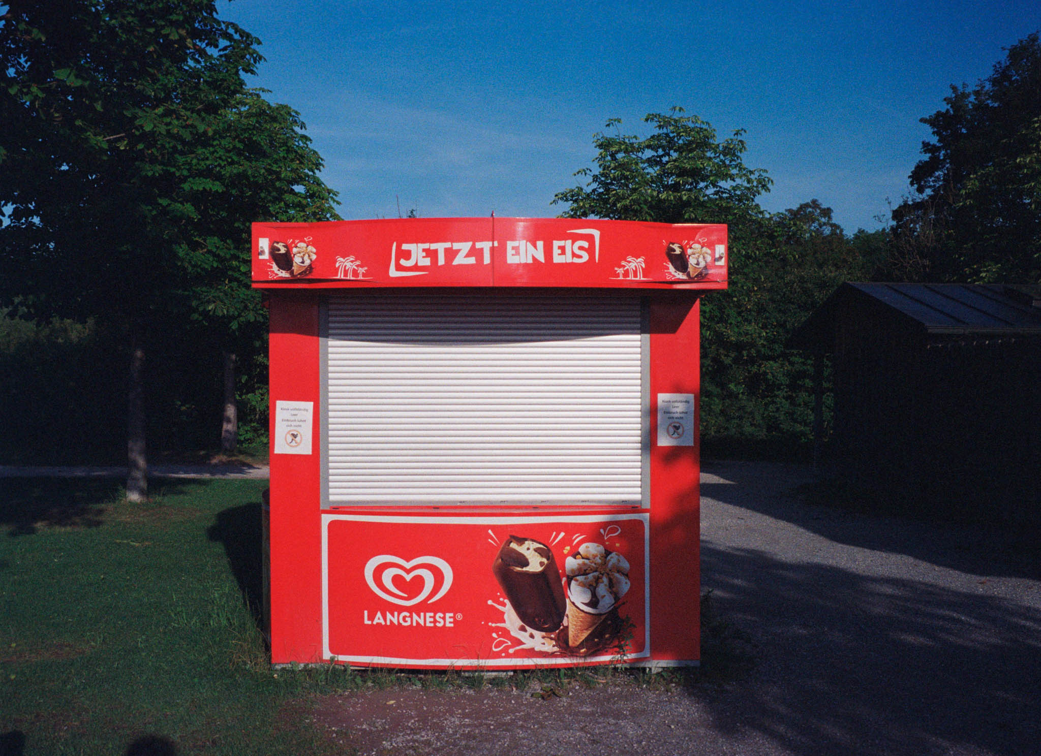 Red ice cream kiosk with a closed shutter, featuring Langnese logo and images of chocolate and vanilla ice cream.