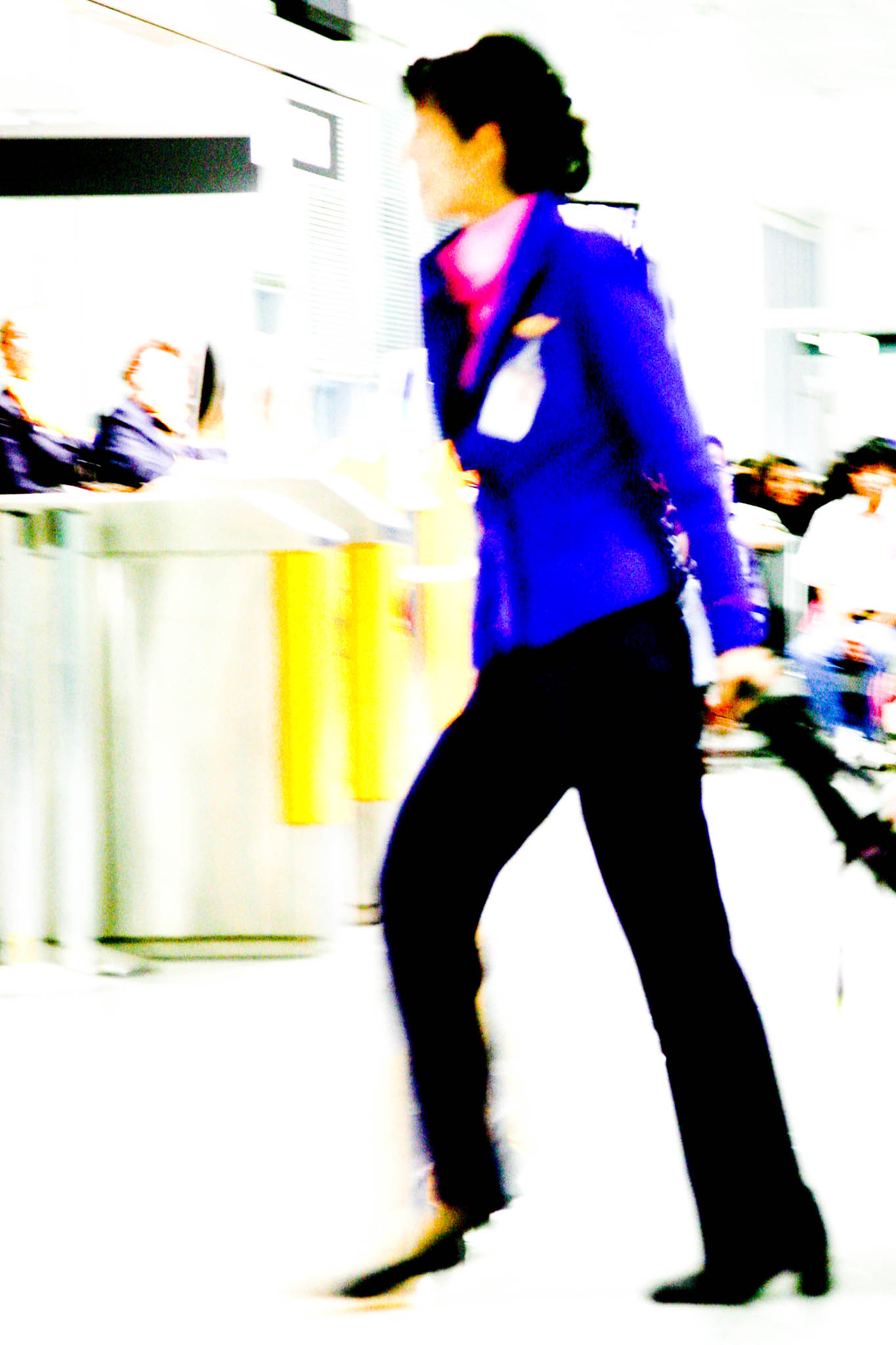 Blurry image of a woman in a blue uniform walking quickly through an airport terminal, pulling a suitcase.