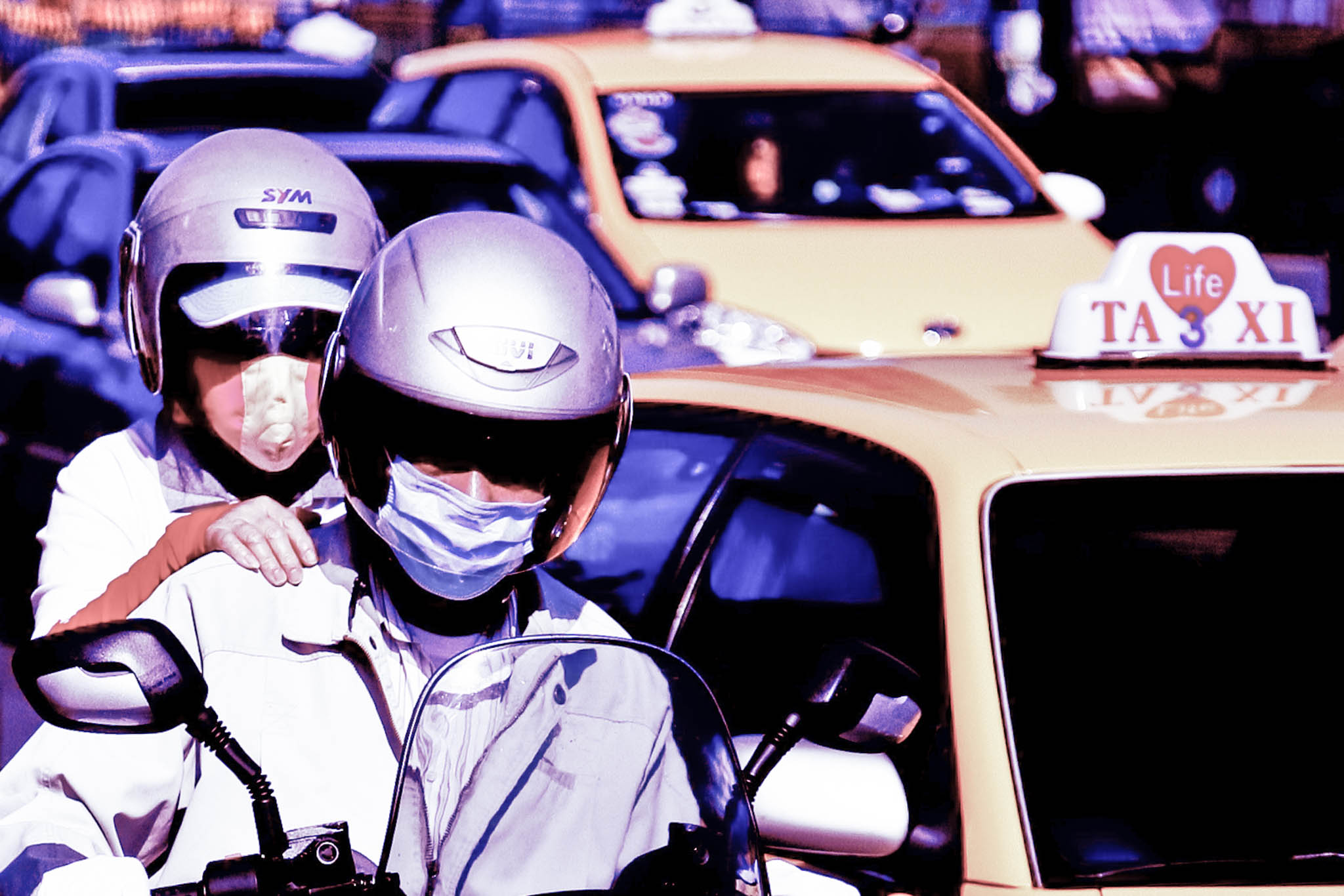 Two people on a motorbike wearing helmets and masks, with yellow taxis and traffic in the background.