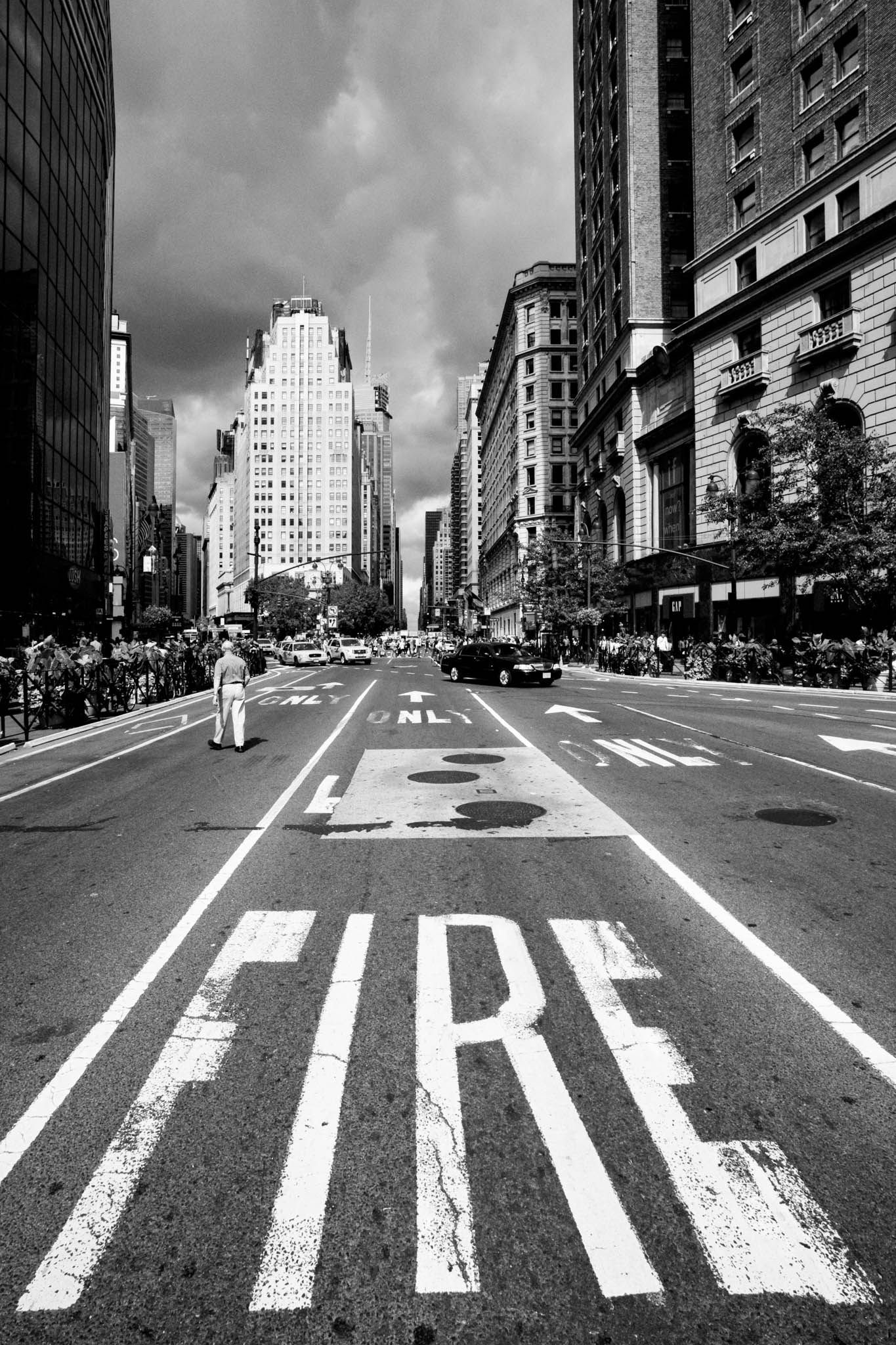 Black and white city street with FIRE painted, tall buildings, and pedestrians crossing under a cloudy sky.