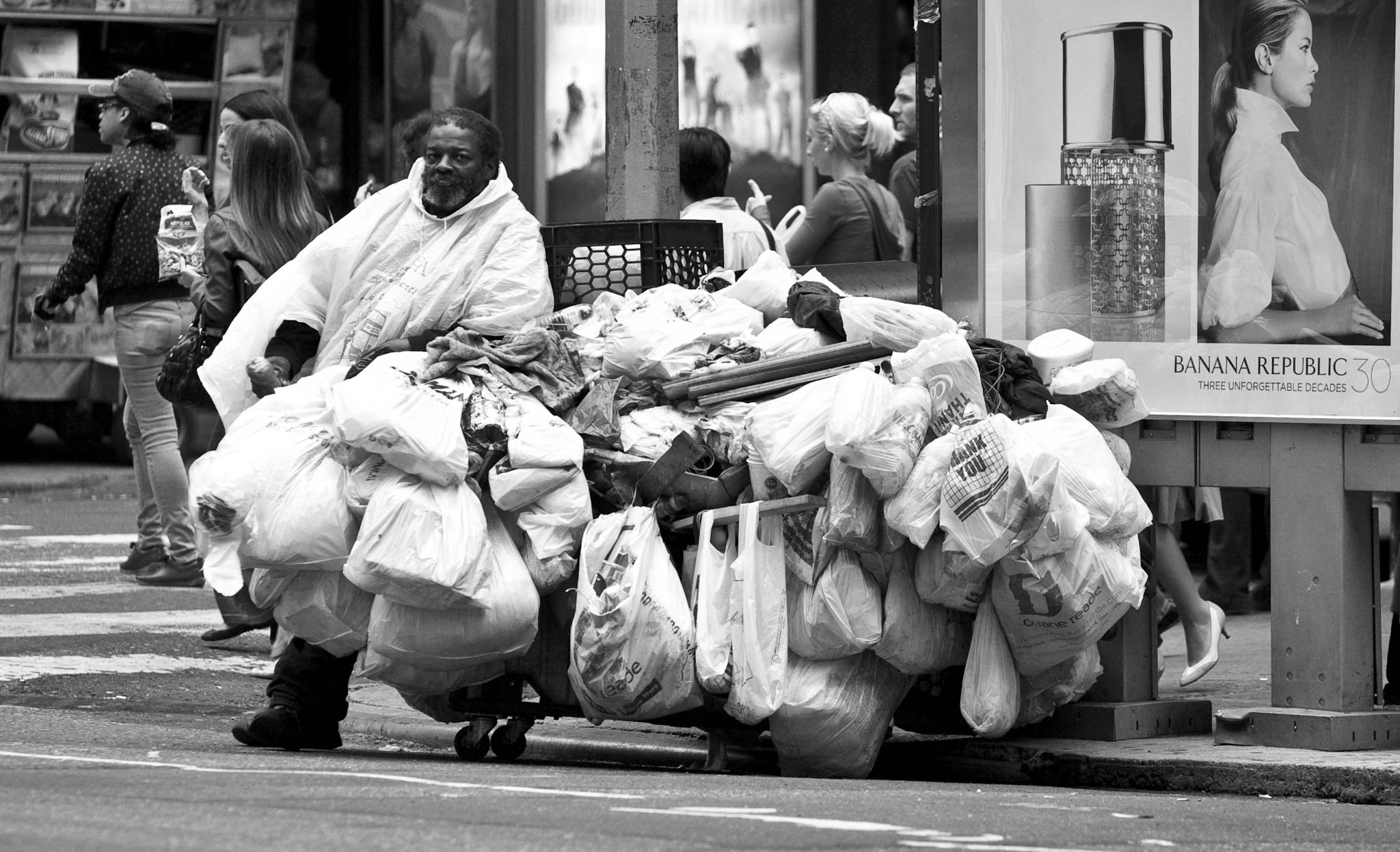 Man with cart full of bags stands on city street near a billboard of a woman; pedestrians walk by.