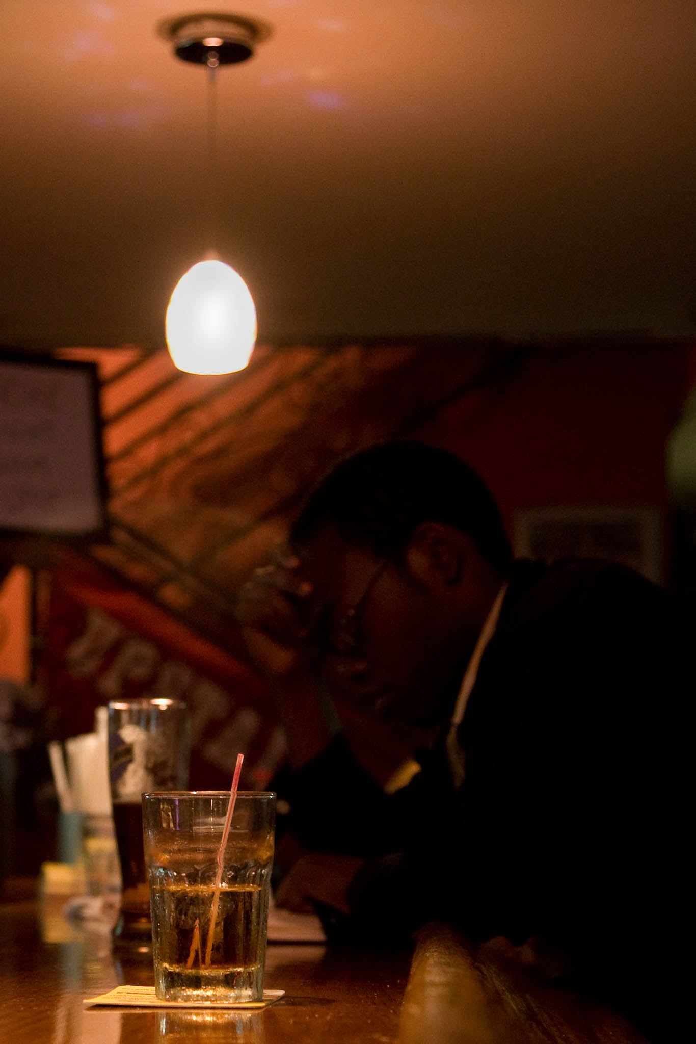 Dimly lit bar with focus on a glass of whiskey; person seated in background under a hanging light.
