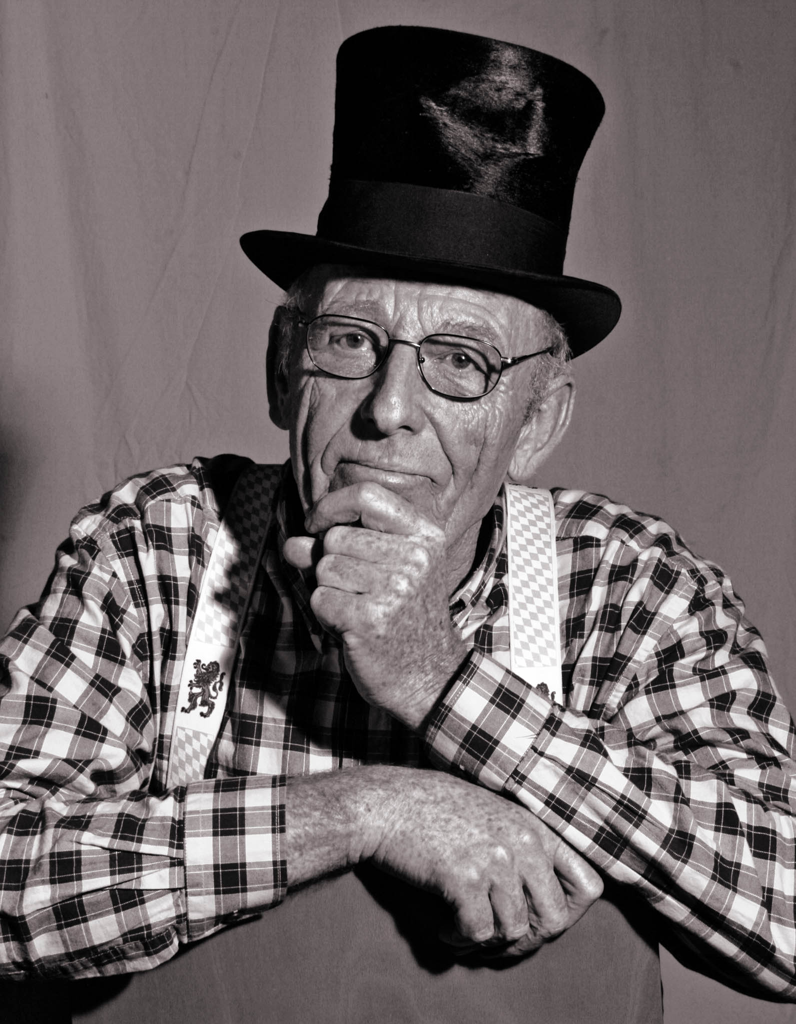 Elderly man in checkered shirt and top hat poses pensively, resting chin on hand against a plain backdrop.