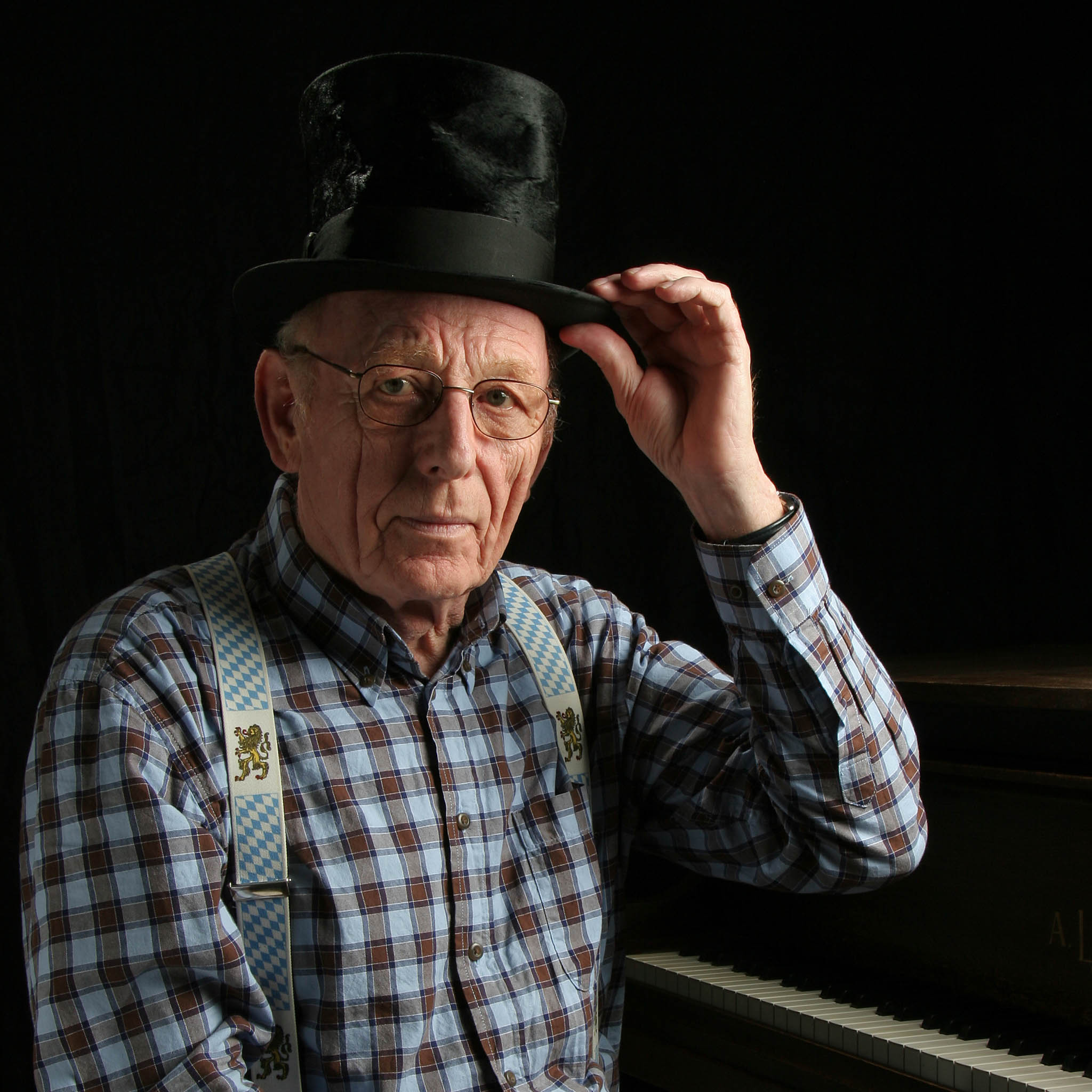 Elderly man in a plaid shirt and top hat, sitting at a piano, touching his hat brim.