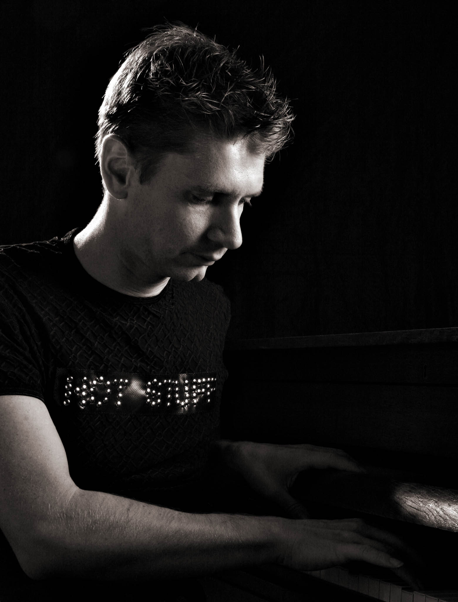 Man in dark shirt plays piano in soft, low-key lighting, expression focused and calm.