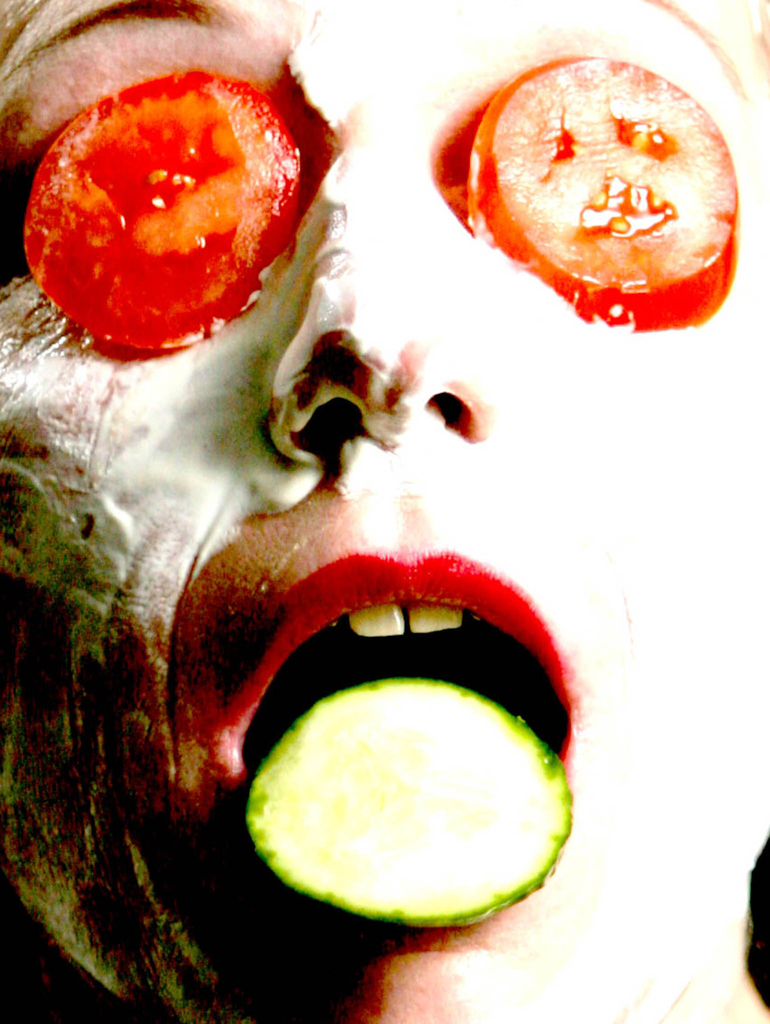 Face covered in white cream, with tomatoes on eyes and cucumber in mouth, depicting a humorous spa treatment scene.