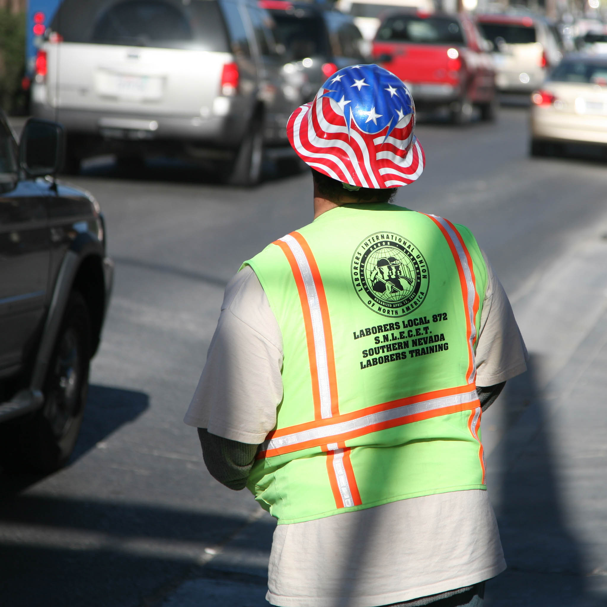 Construction worker in a bright safety vest and American flag hard hat on a busy street for laborer training.