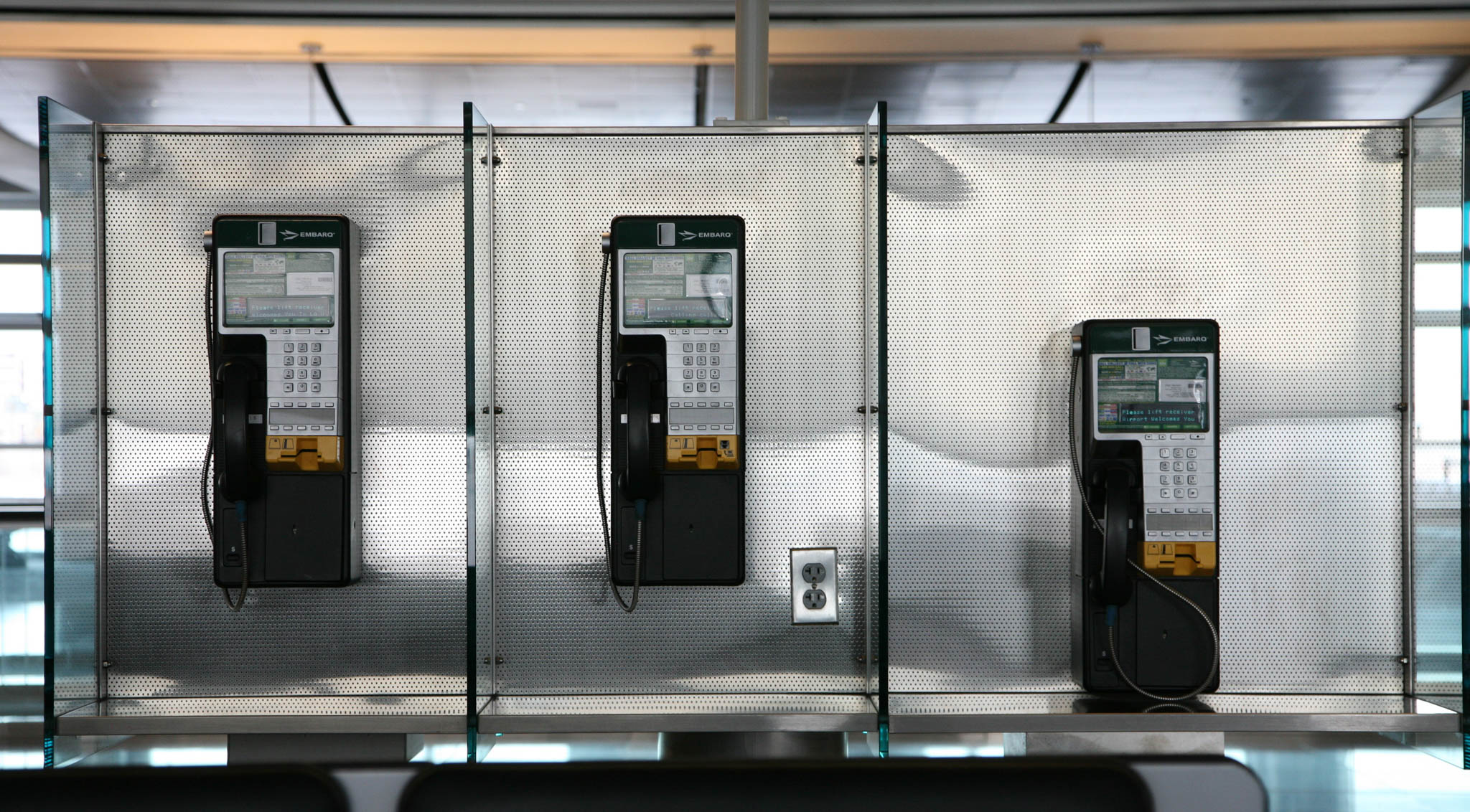 Three modern payphones with digital screens mounted on a metal wall, featuring a power outlet below.