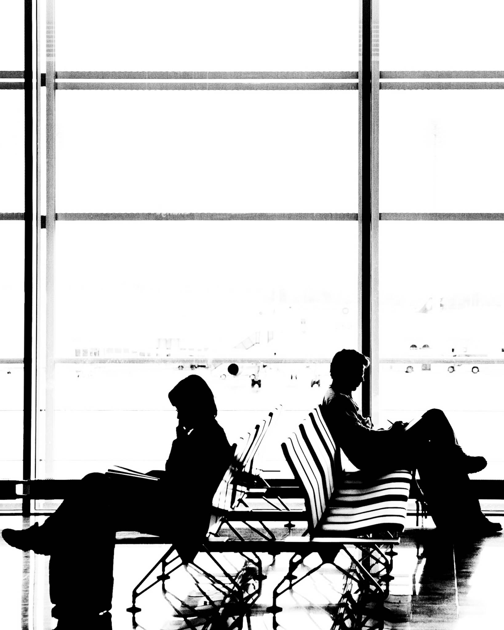 Silhouetted travelers sit back-to-back in an airport terminal, framed by large windows and an airplane in the background.