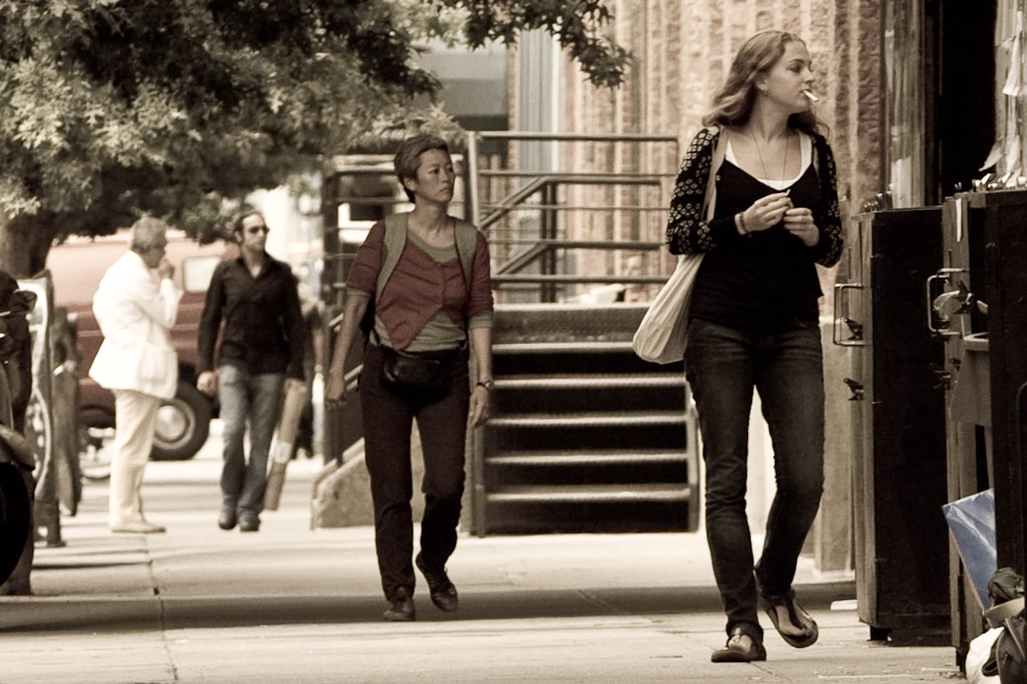 People walking on a city sidewalk under trees, woman in front holds a cigarette and wears a patterned sweater.