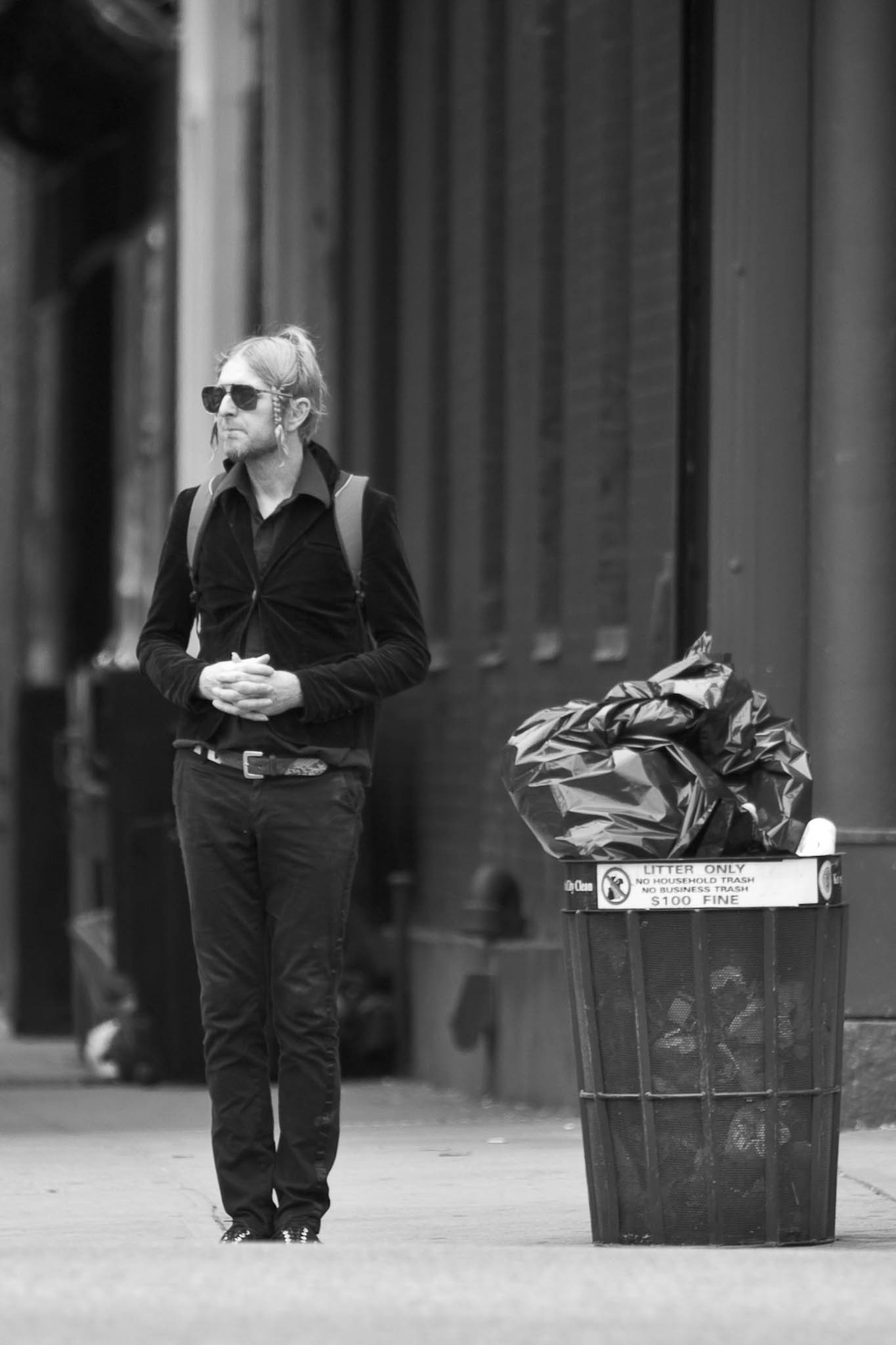 Man in black attire stands near a trash can on a city street, wearing sunglasses and a backpack, in a black and white photo.