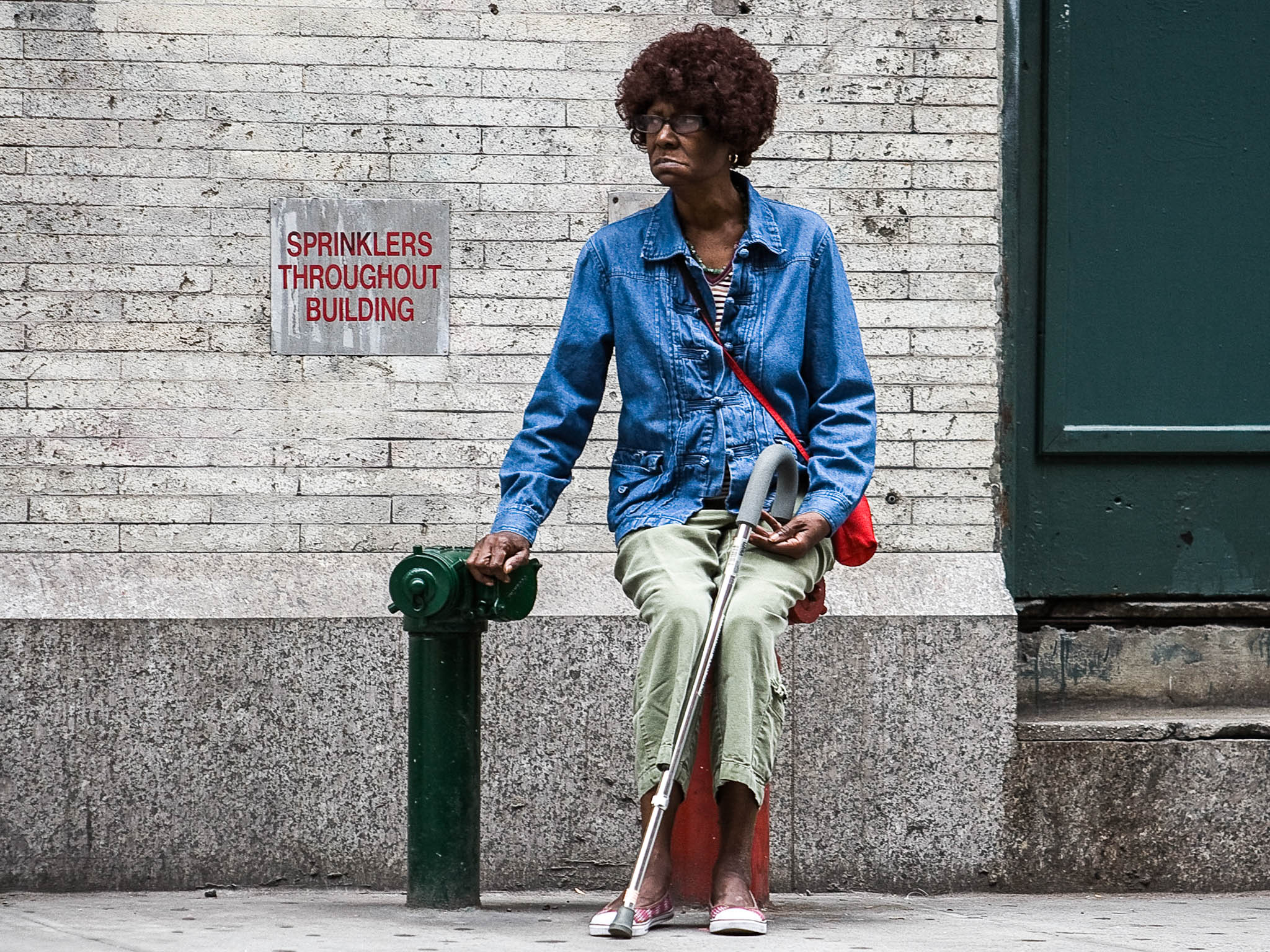Elderly woman with a cane sitting on a street hydrant by a brick wall in casual attire.